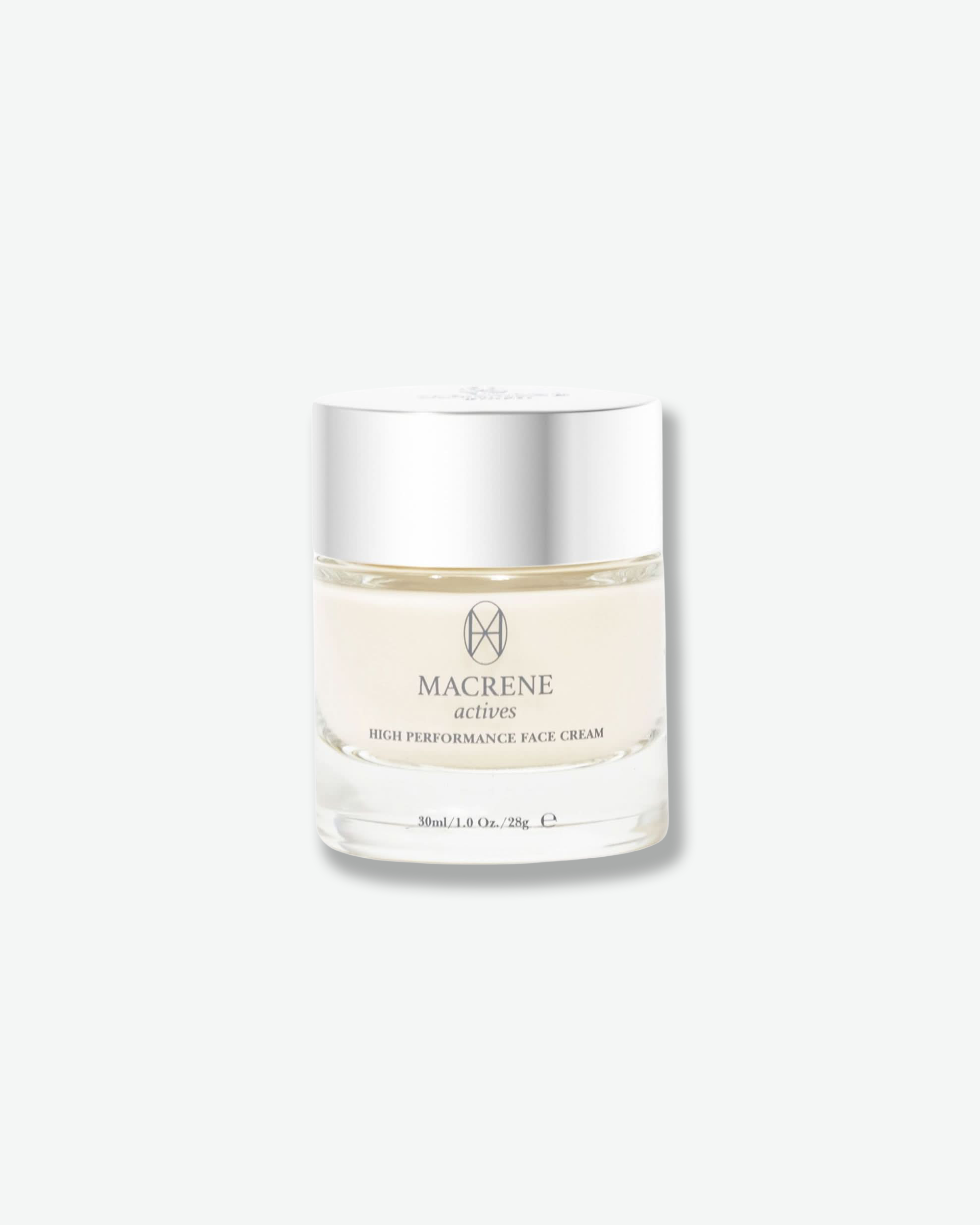 Front view of a Macrene Actives High Performance Face Cream jar against a plain gray background. The premium container features a heavy, clear glass base showcasing the off-white cream inside, a minimalist dark grey logo and branding text, and a smooth, brushed silver metallic cap.