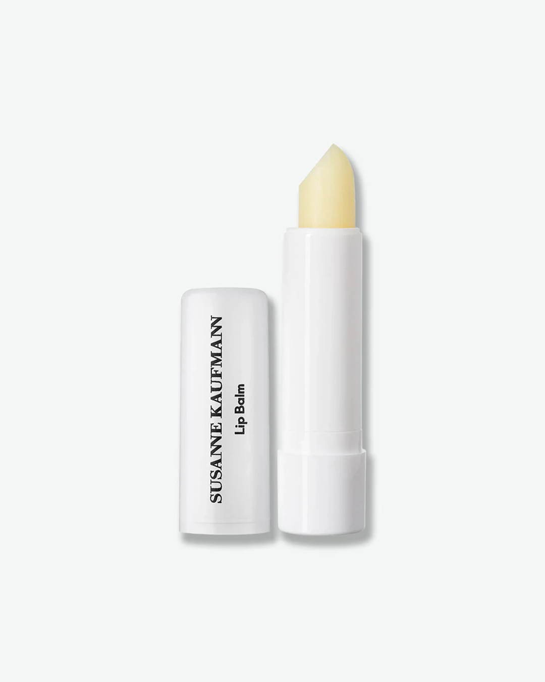 A product shot displaying an open tube of Susanne Kaufmann Lip Balm against a plain light gray background. On the right stands the slim, white plastic lipstick-style tube, rolled up to reveal a smooth, angled pale yellow lip balm stick. On the left, the matching white plastic cap rests upright, showing the brand name "SUSANNE KAUFMANN" and "Lip Balm" printed vertically in black.