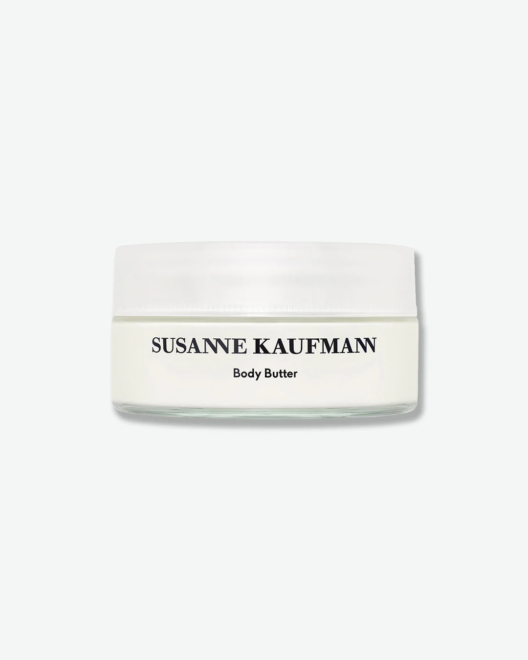 A product shot of a Susanne Kaufmann Body Butter jar, centered against a plain light gray background. The shallow clear glass jar is filled with a smooth, solid white cream formula and is topped with a flush, frosted white screw cap. The front displays the brand name "SUSANNE KAUFMANN" and the product title "Body Butter" in minimalist black typography.