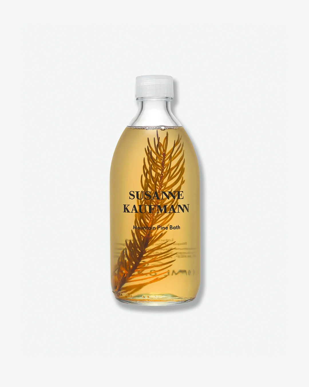 A product shot of a Susanne Kaufmann Mountain Pine Bath bottle, centered against a plain light gray background. Inside the curved clear glass bottle, a large, dark green pine sprig branch is completely submerged within a translucent, light amber liquid formula. The bottle is capped with a clear plastic screw top and features black branding text on the front.