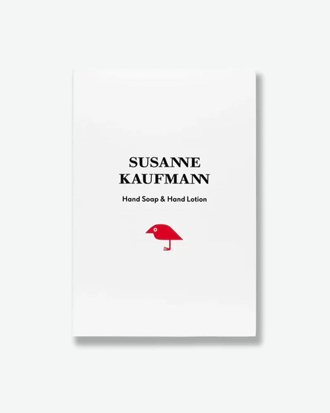 A product shot of the white cardboard packaging box for a Susanne Kaufmann Hand Soap & Hand Lotion set, centered against a plain light gray background. The minimalist white box features the brand name in black serif typography above the product name. Near the bottom is a single, graphic illustration of a red bird silhouette standing on one leg.