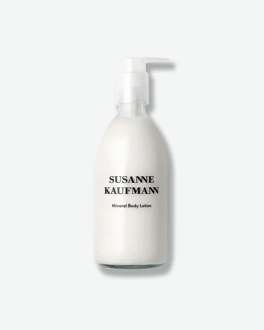 A product shot of a Susanne Kaufmann Mineral Body Lotion bottle, centered against a plain light gray background. The curved bottle is crafted from frosted clear glass, showing a solid white cream lotion inside. It features a clear plastic pump mechanism and clean black typography on the front.