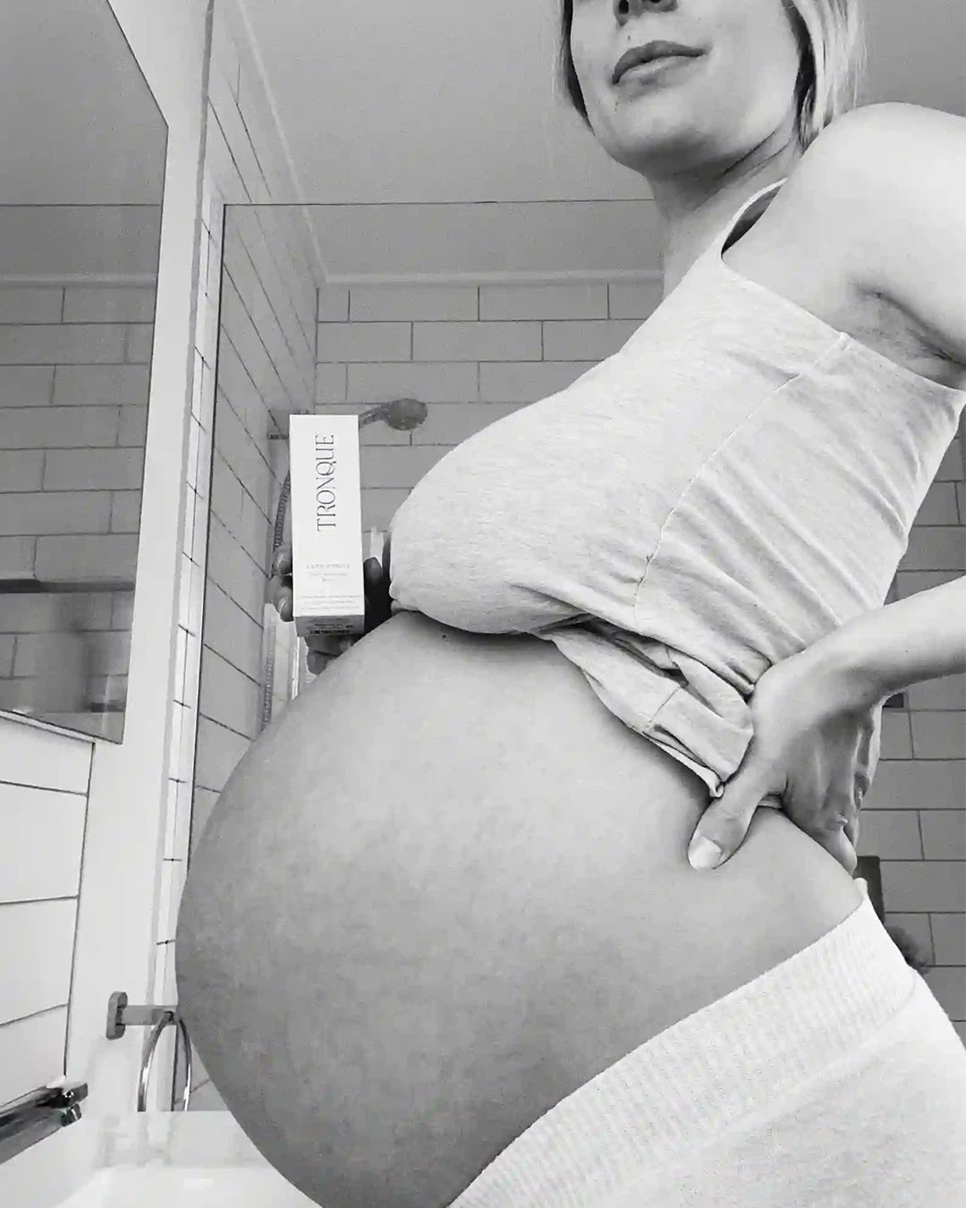 Black and white photo of a pregnant person holding a box labeled 'Tronque' in a bathroom.