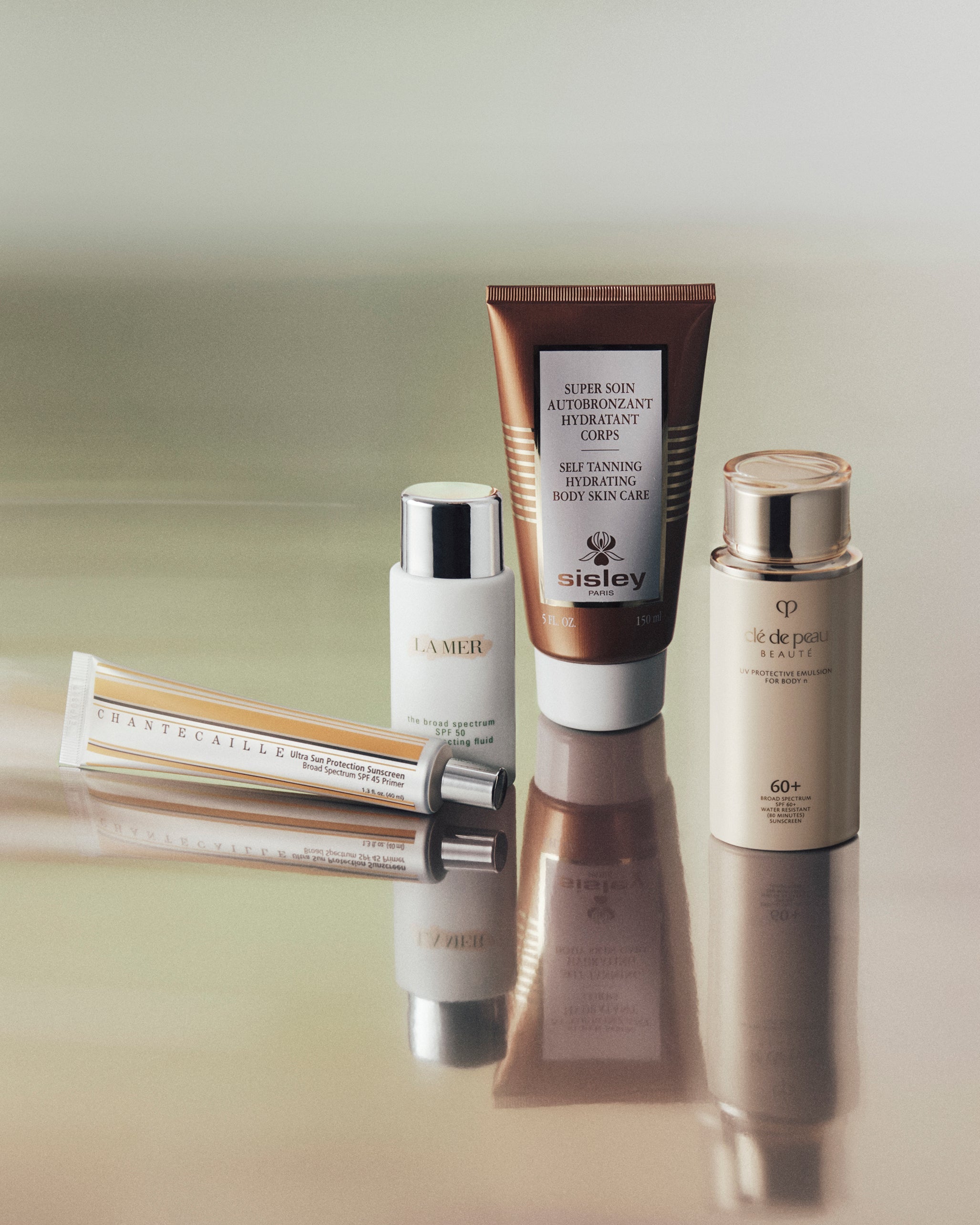 Four skincare products arranged on a reflective surface against a soft beige background: a brown tube of aisley Super Rich Cream, a white bottle of La Mer Advanced Night Repair, a beige bottle of TF de Coeur Ultra-Pure Moisturizing Lotion, and a rolled cream tube with metallic detailing.