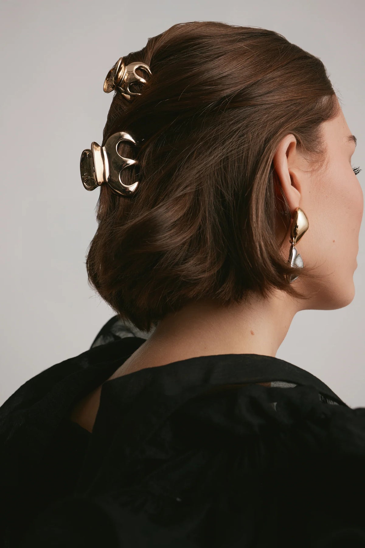 Woman with glossy brunette hair styled half-up with two sculptural gold claw clips, wearing statement drop earrings and a black top.