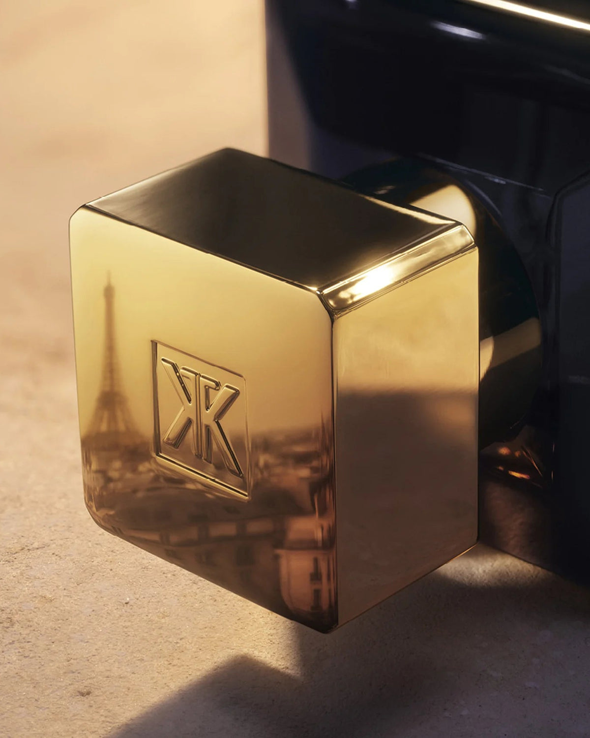A close-up of a polished gold cube-shaped accessory with an embossed logo, resting against a dark glossy surface on a warm beige surface.