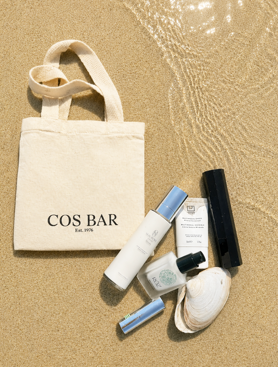 A beige canvas tote bag with 'COS BAR Est. 1975' printed on the front, arranged on sandy beach ground alongside several COS BAR skincare bottles and a white clam shell.