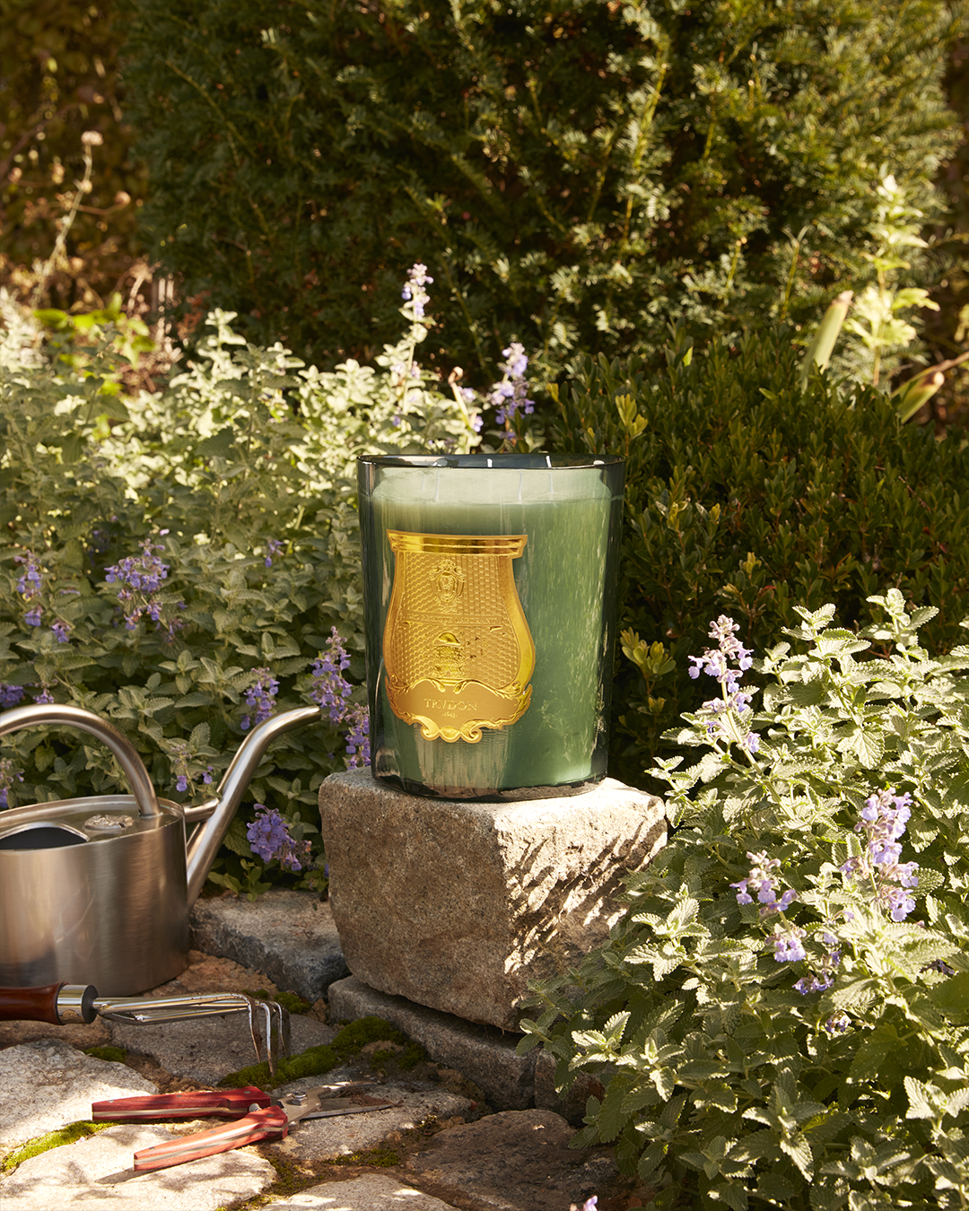 A large green Trudon scented candle resting on a stone block in a garden surrounded by green plants and purple flowers.