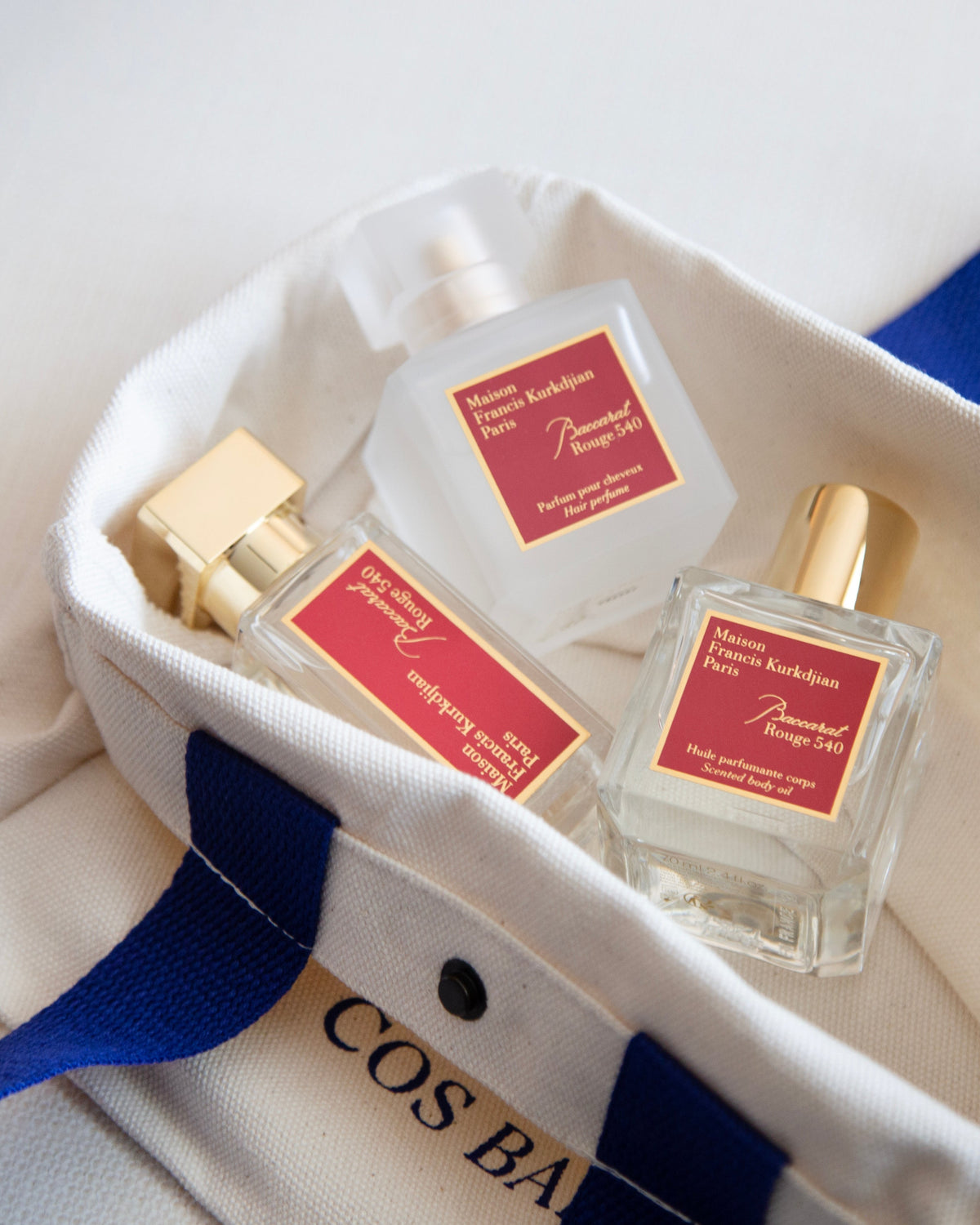 Three small perfume bottles with red labels and gold caps arranged inside a white tote bag with blue handles. The tote bag has partial gold lettering reading 'COS BAR'