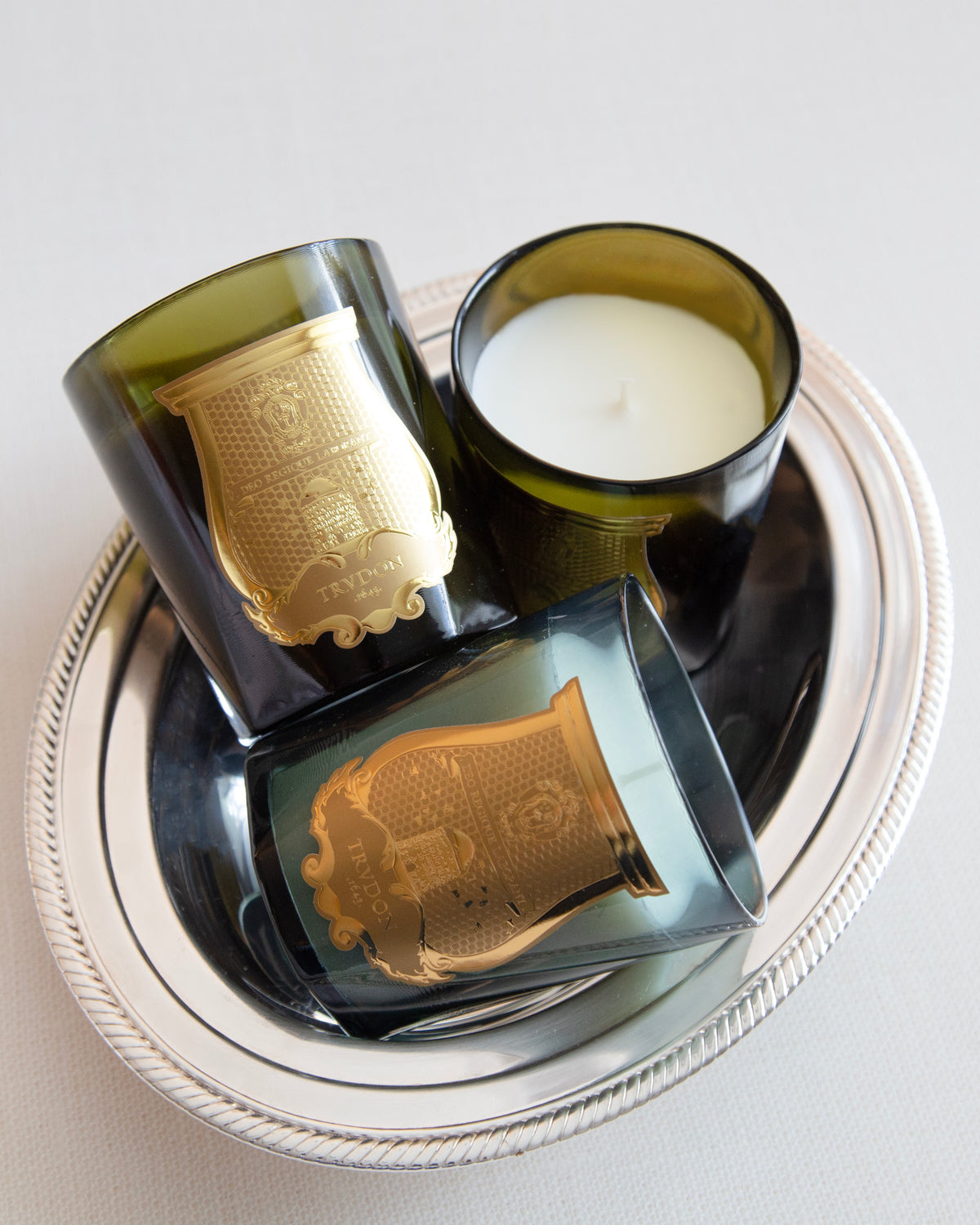 Three Trudon candles in dark green glass vessels with ornate gold emblems, arranged on a silver tray.