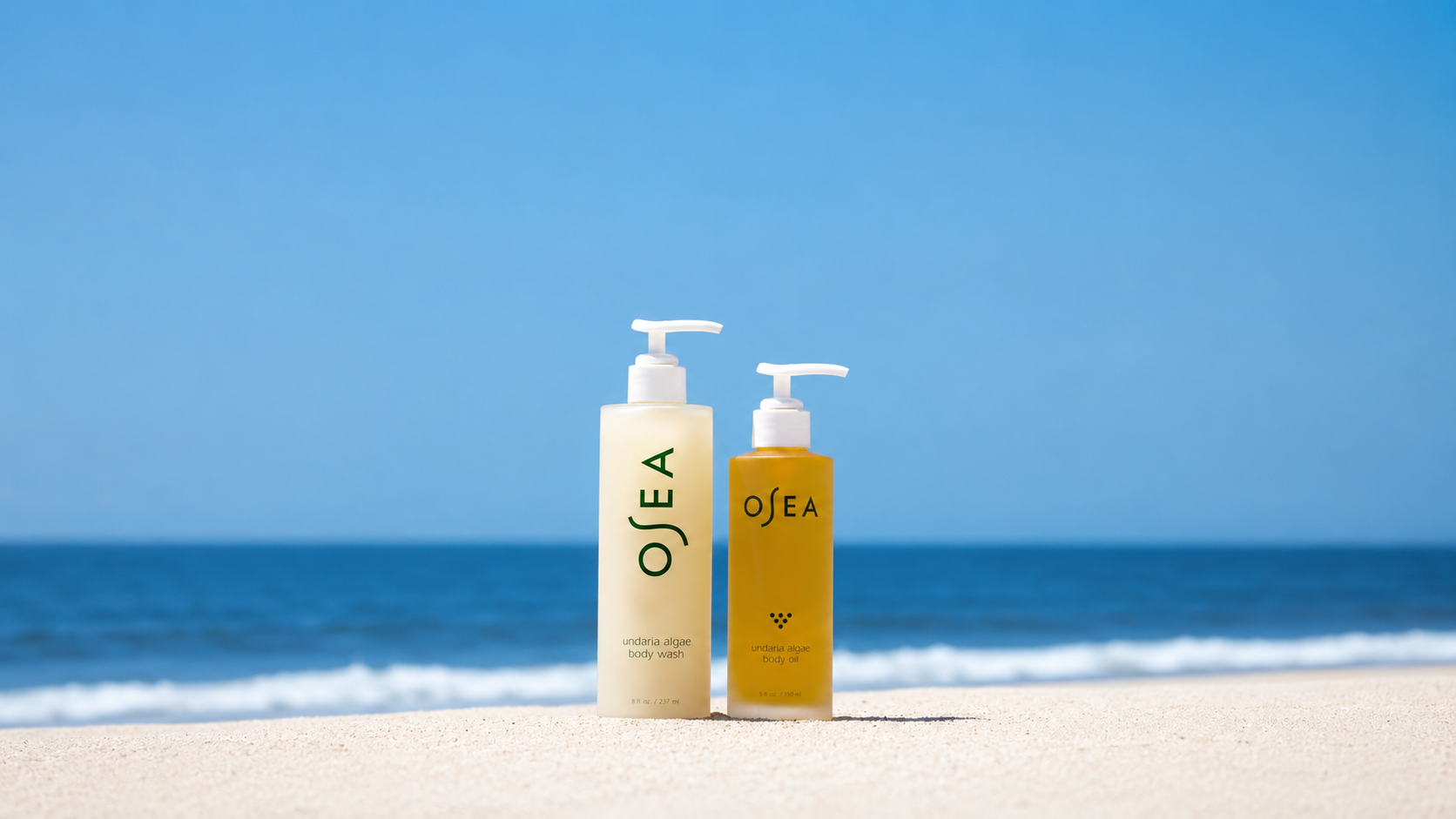 Two OSEA Undaria Algae body care products—a cream-colored Body Wash and an amber Body Oil with white pumps—stand on soft white sand against a bright blue ocean and clear sky. The minimalist beach scene evokes a fresh, sunlit coastal atmosphere.