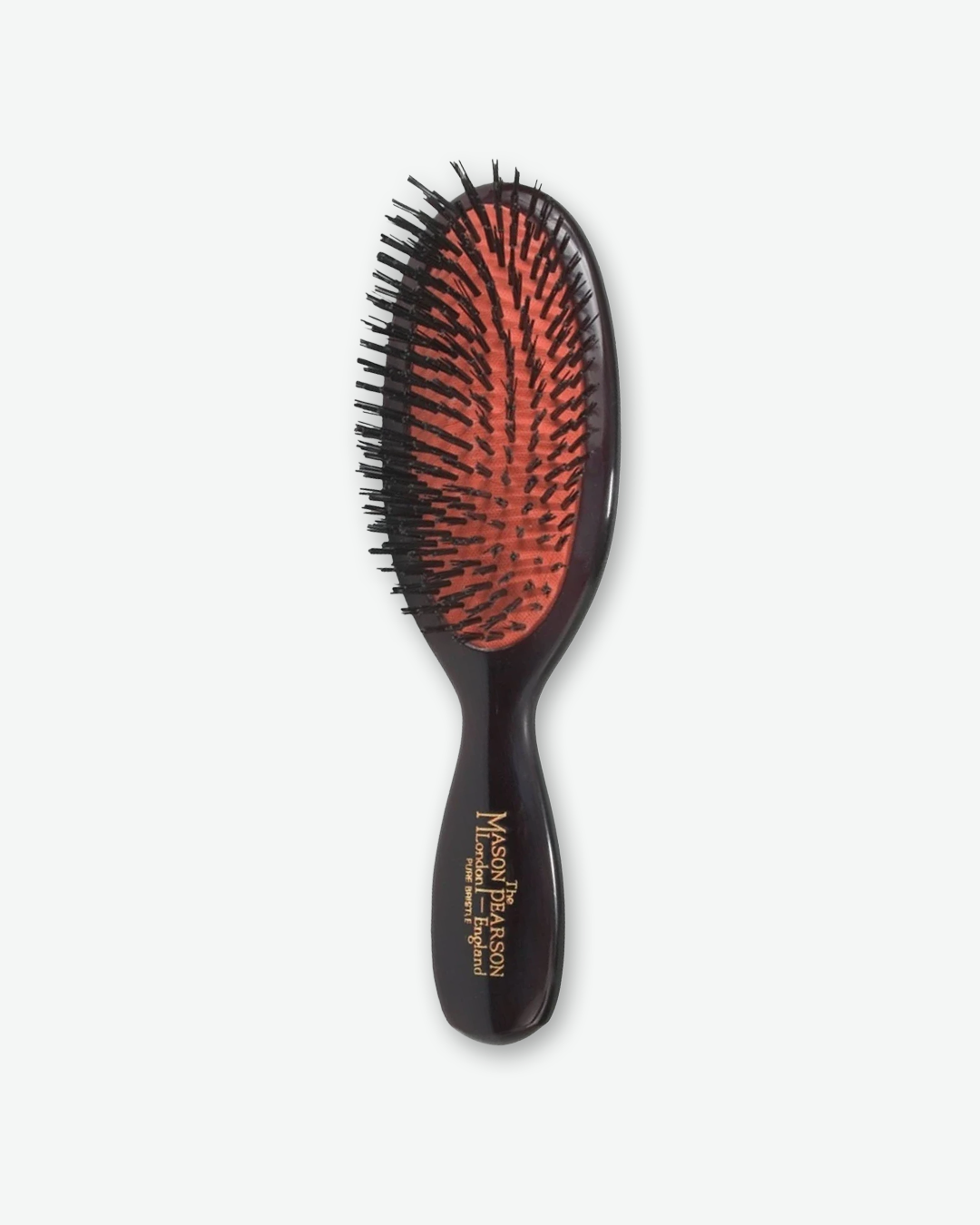 Front view of a small Mason Pearson pocket-sized oval hairbrush in dark ruby, featuring a bright orange rubber cushion, black pure boar bristles, and gold text on the handle that reads "The Mason Pearson London – England Pure Bristle".