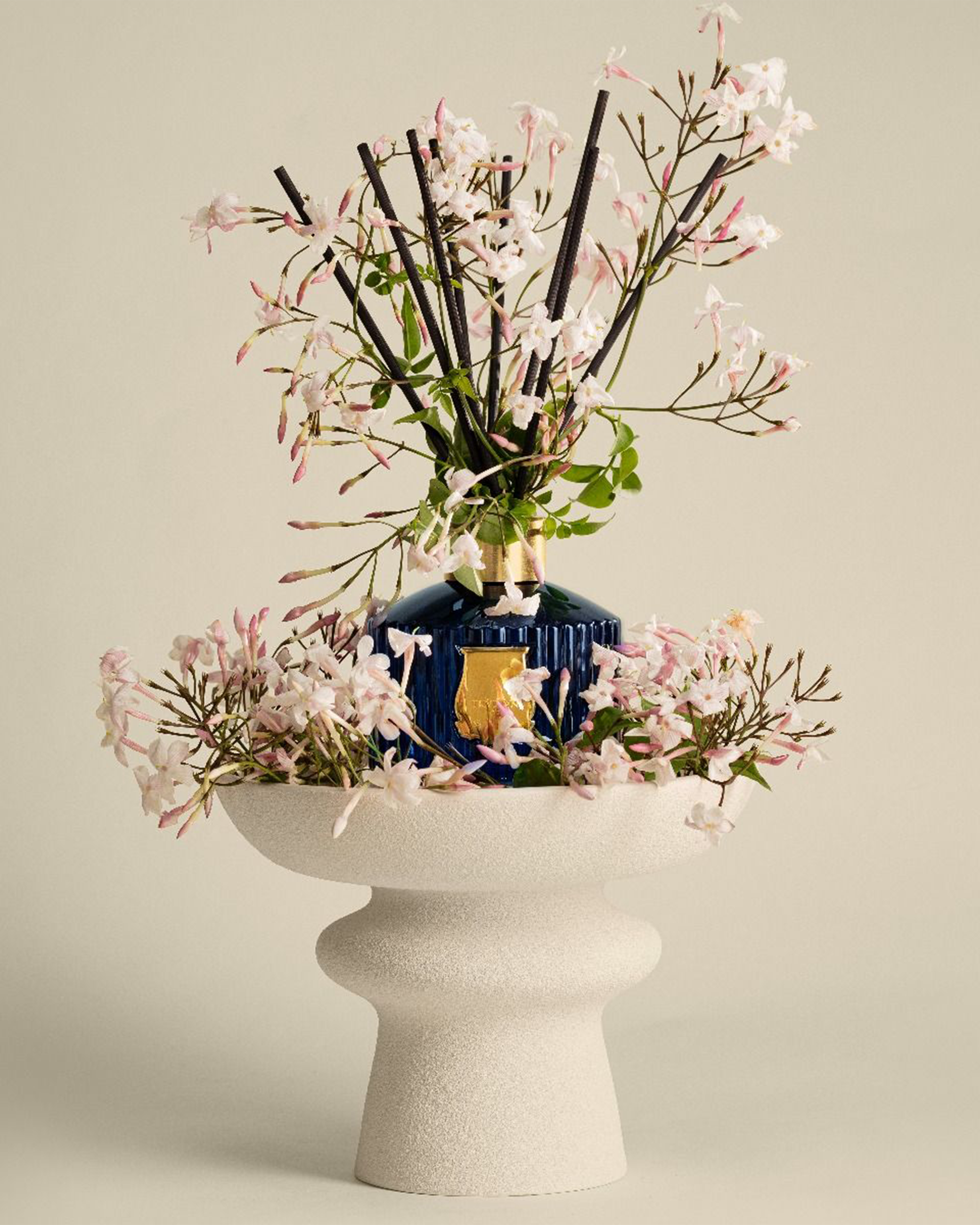 A decorative blue glass perfume bottle with gold detailing, surrounded by pale pink blossoms and dark reed sticks, displayed in a textured white pedestal bowl against a warm beige background.