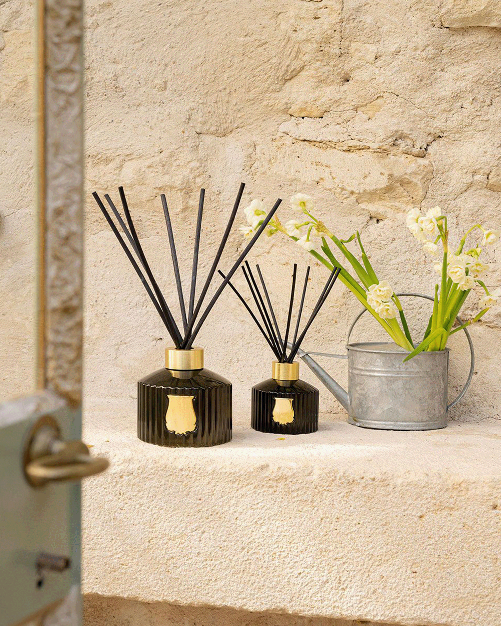 Two black reed diffusers with gold caps sit on a stone ledge next to a galvanized metal watering can holding white flowers, against a textured beige stone wall.