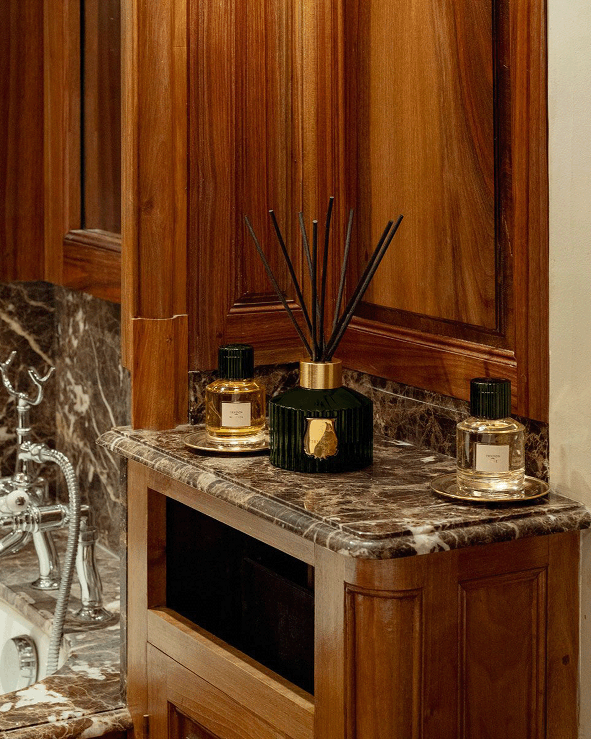 A luxury reed diffuser set displayed on a dark marble bathroom vanity. The set includes a central ribbed dark green reed diffuser with gold accents and three matching amber glass bottles with gold caps and labels.