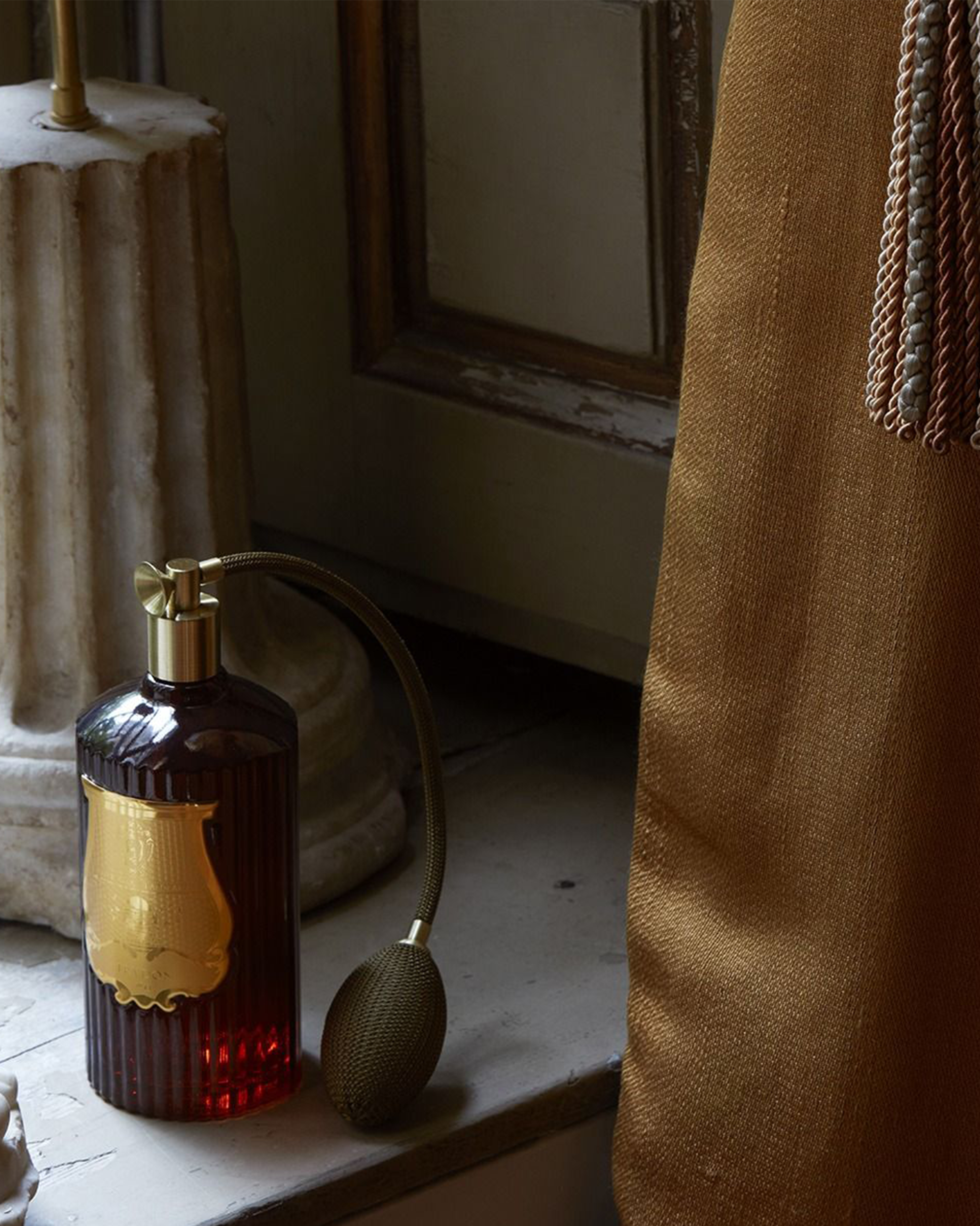 A vintage-style amber glass perfume bottle with a gold label and pump, connected to a bulb pump sprayer, sitting on a windowsill next to a brown curtain.