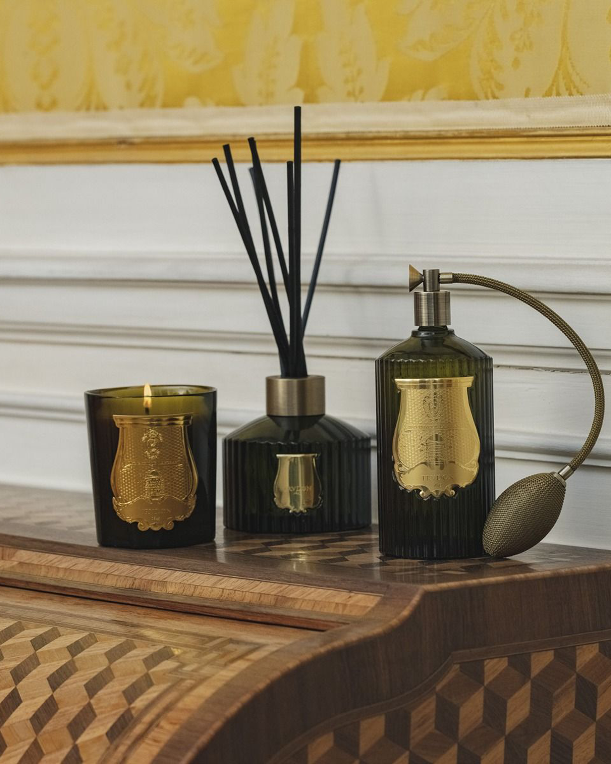 Three dark green glass home fragrance items with gold labels are arranged on a wooden table against a white paneled wall and yellow patterned wallpaper. The set includes a candle, a reed diffuser with black reeds, and a perfume-style atomizer with a curved metal sprayer.