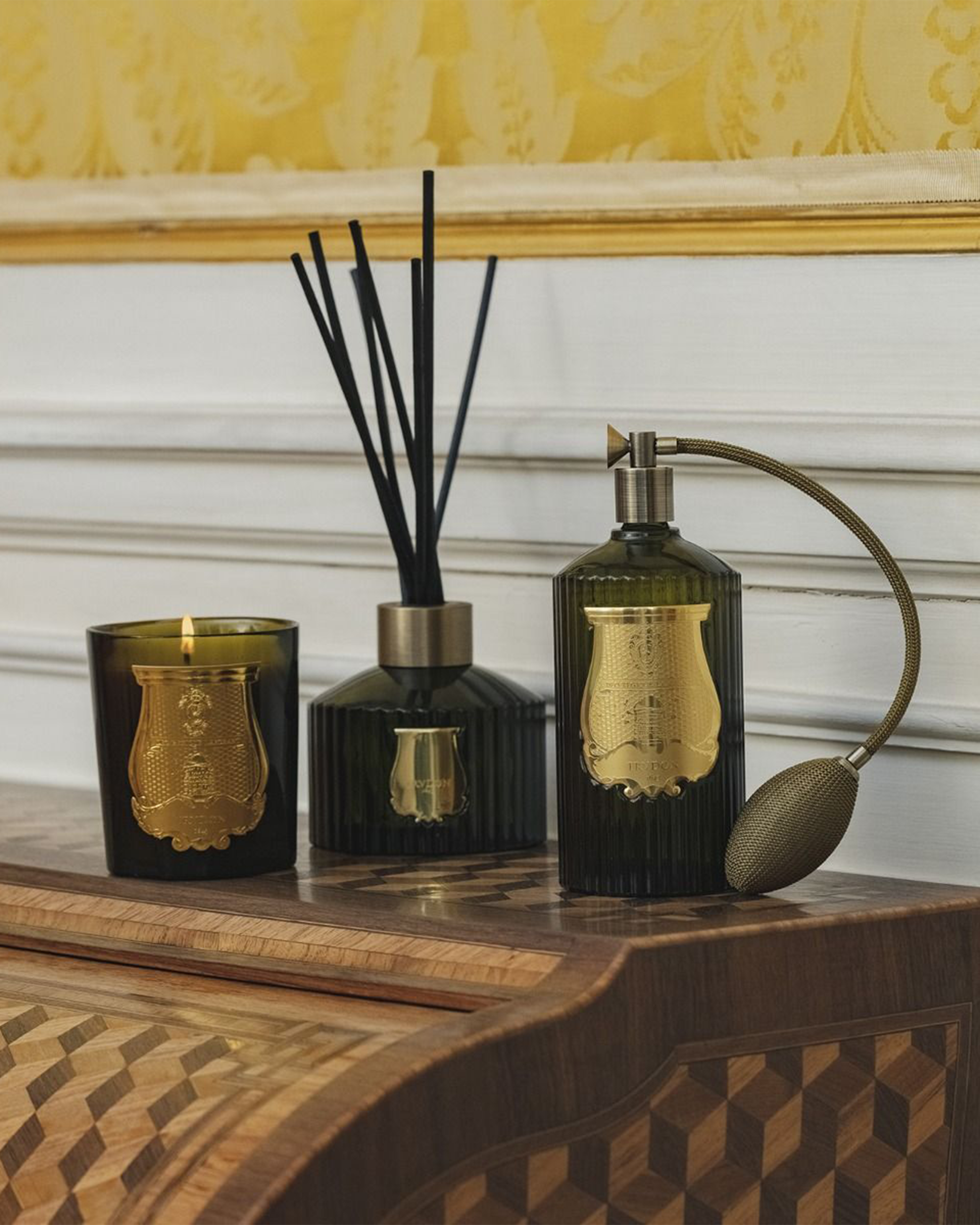 Three luxury home fragrance items in dark green glass with gold embossed labels: a candle, a reed diffuser, and a perfume atomizer, arranged on a wooden console table against a white paneled wall and gold wallpaper.
