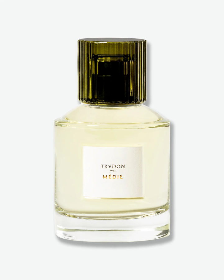 A front-facing studio shot of Trudon Médie Eau de Parfum, featuring a heavy clear glass bottle filled with pale yellow fragrance and topped with a fluted dark olive green cap.