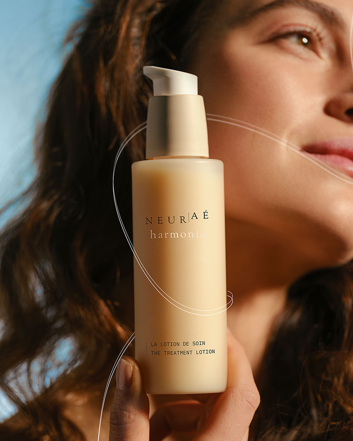 A hand holding the Neuraé Harmonie Treatment Lotion bottle with a smiling woman in the soft-focus background of a warm lifestyle photo.