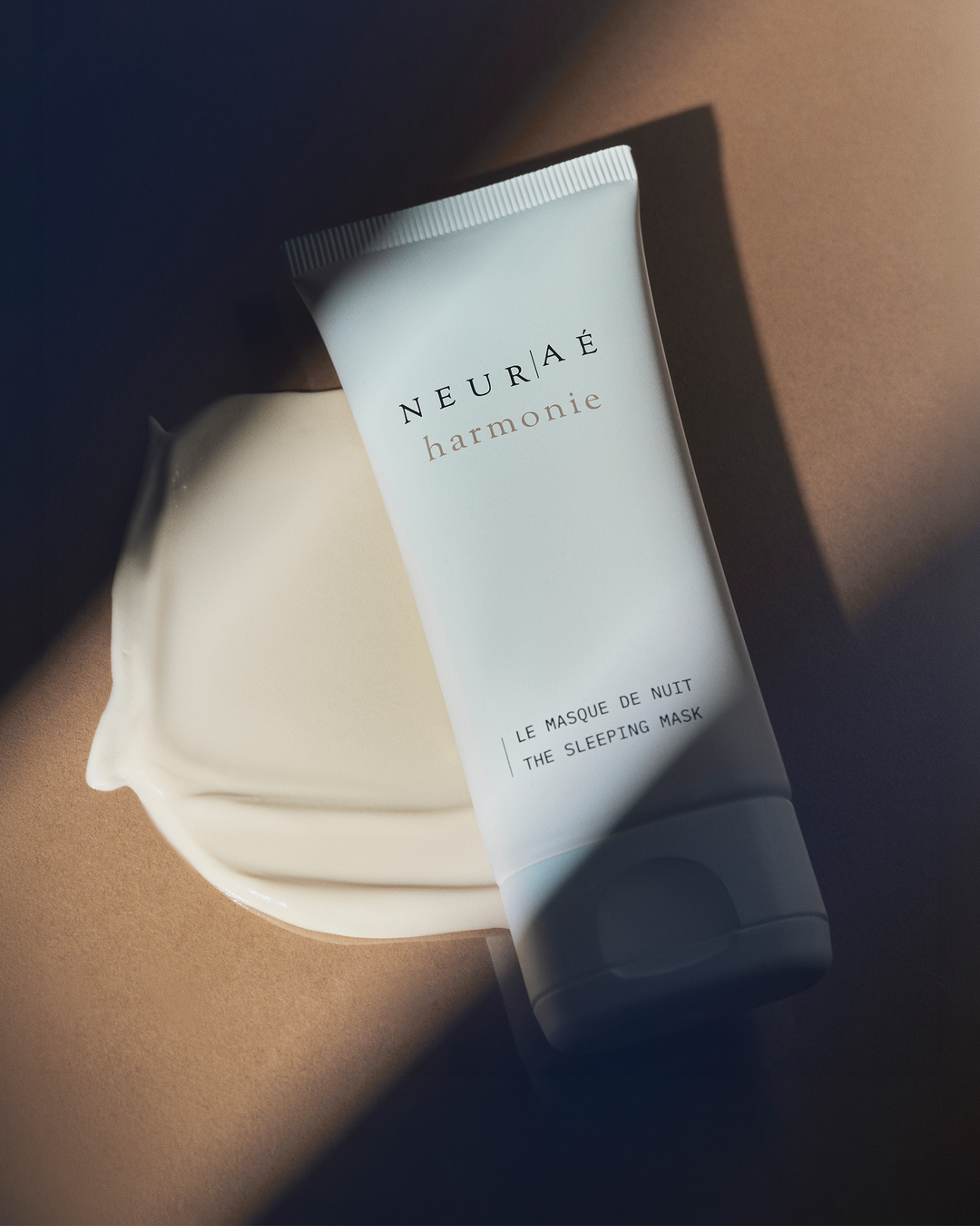 Neurae Harmonie The Sleeping Mask white tube resting diagonally on a smooth smear of its off-white cream texture under dramatic lighting.