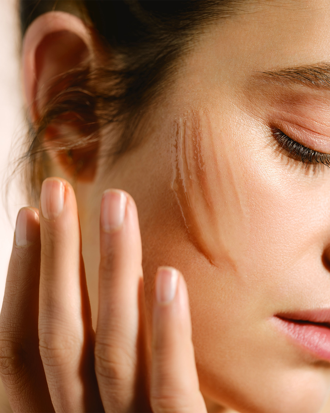 A close-up lifestyle portrait of a person showing a clear gel skincare product swiped across their cheekbone under warm, bright lighting.