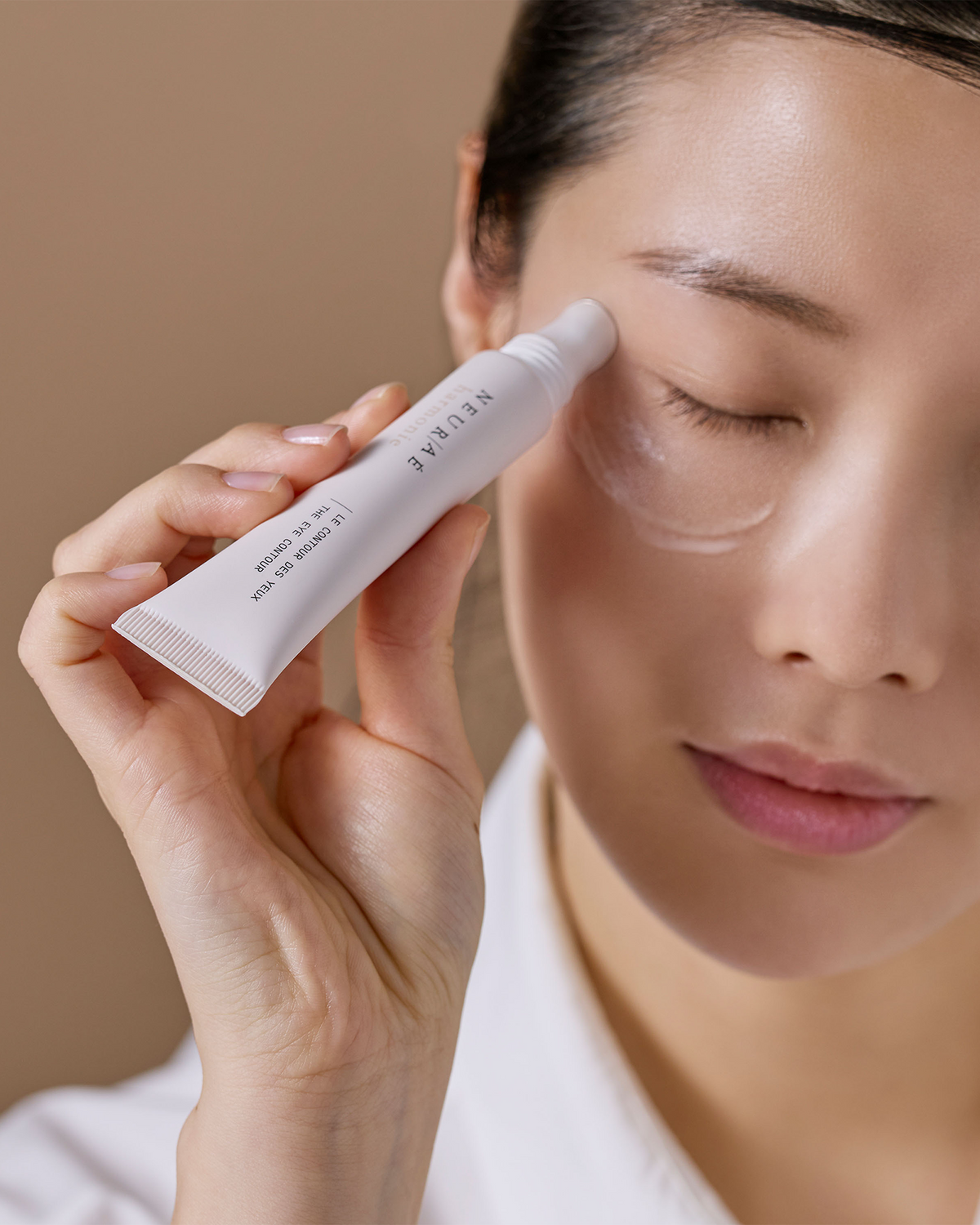A close-up of a woman applying a clear gel cream from a white squeeze tube labeled 'NEURAE' to her eye area, with a soft beige background.