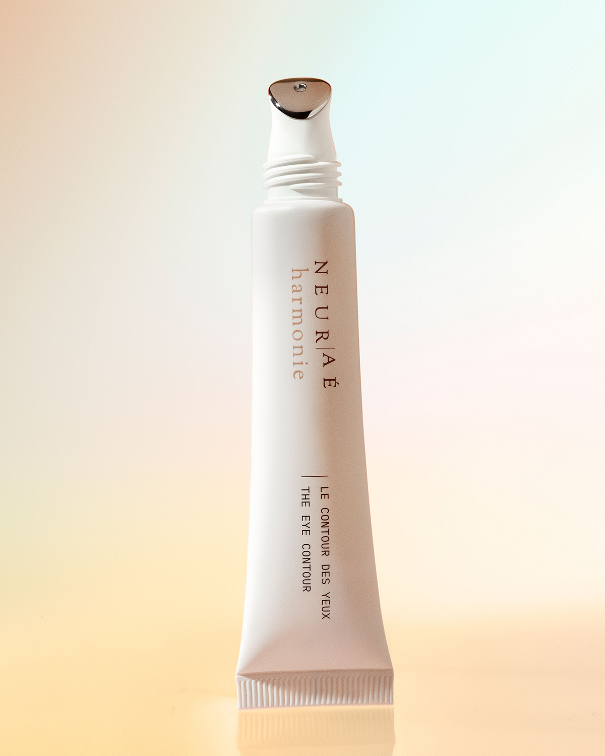A white squeeze tube of NEURAE Harmonie skin cream with a metallic gold cap, standing upright on a reflective surface against a soft pastel gradient background.