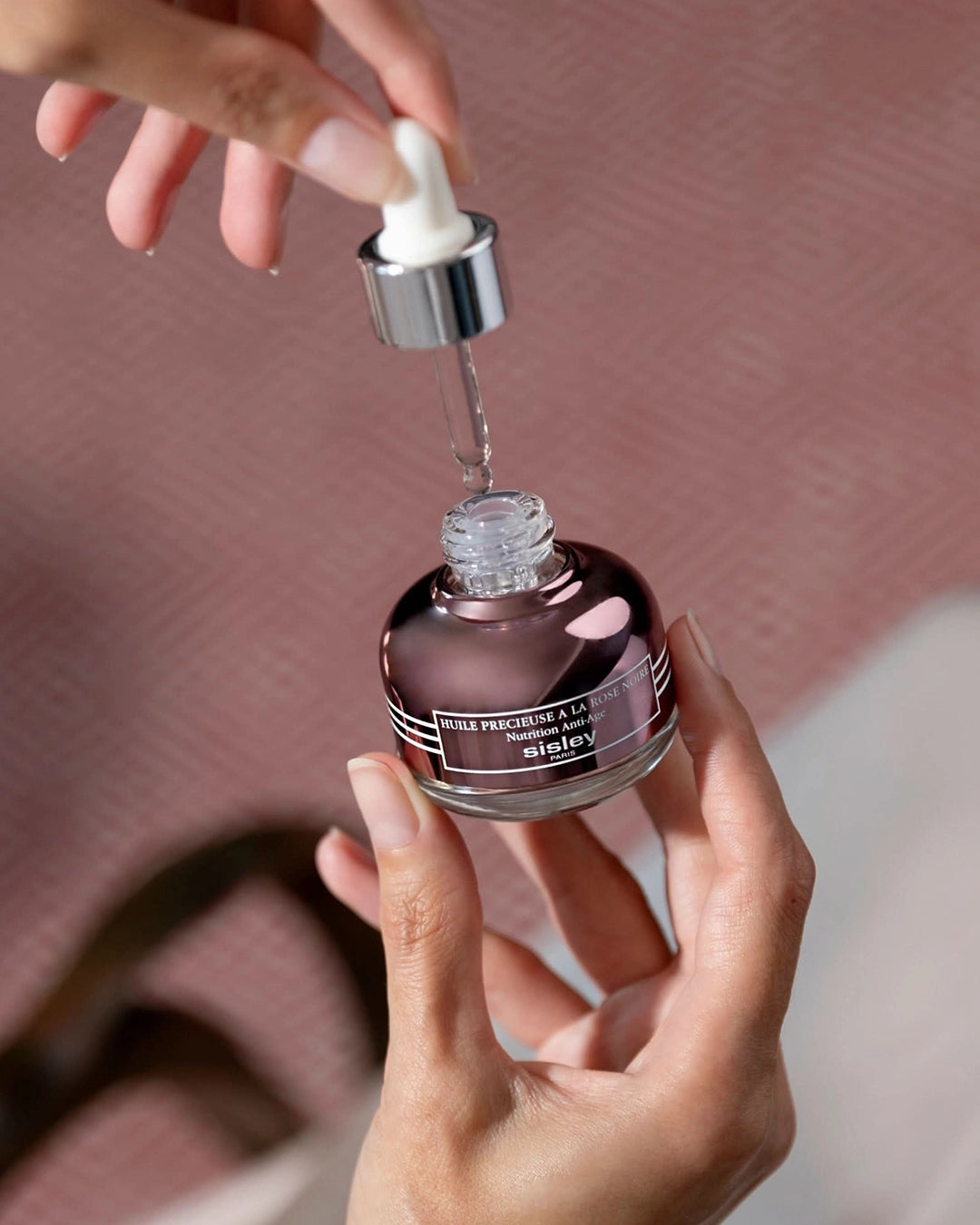 Hand holding a dropper over a bottle of Sisley product with a blurred background