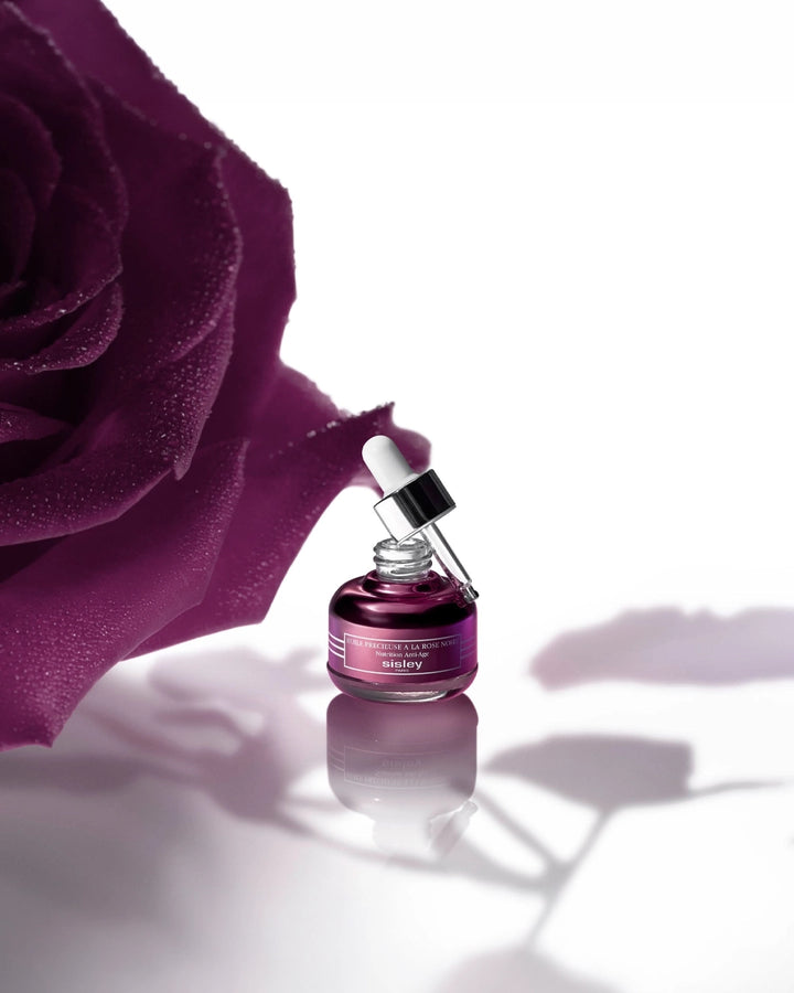 Purple skincare serum bottle with dropper next to a purple rose on a white background