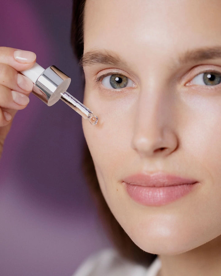 Person applying a dropper of skincare product to their face against a blurred background