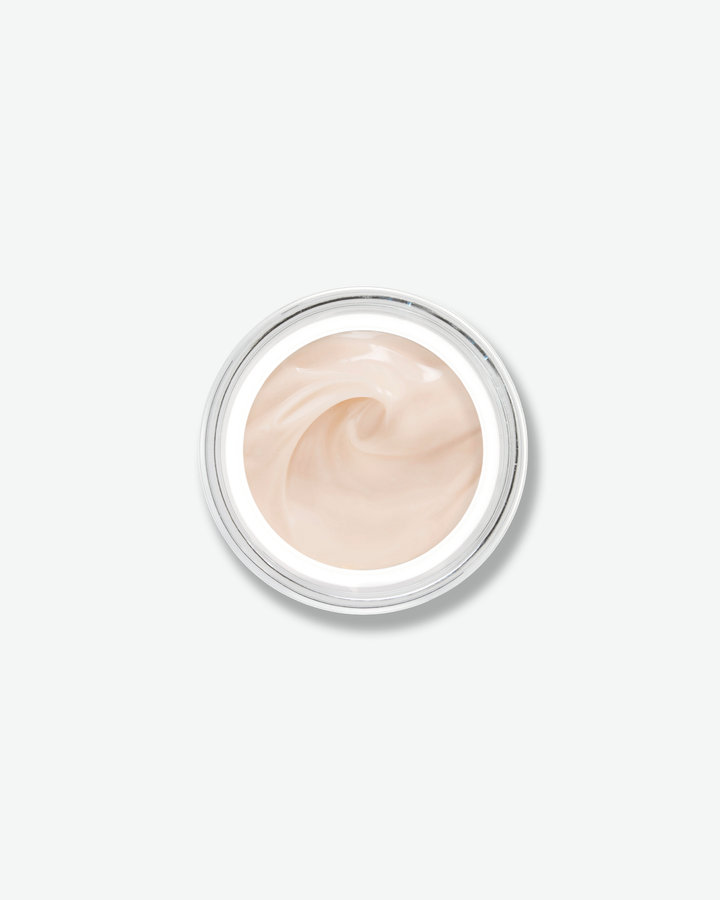 Overhead, top-down view looking directly into an open skincare jar against a plain gray background. The smooth, light-colored glass rim encircles a soft, pale peach-tinted face cream that is textured with an elegant swirl in the center.