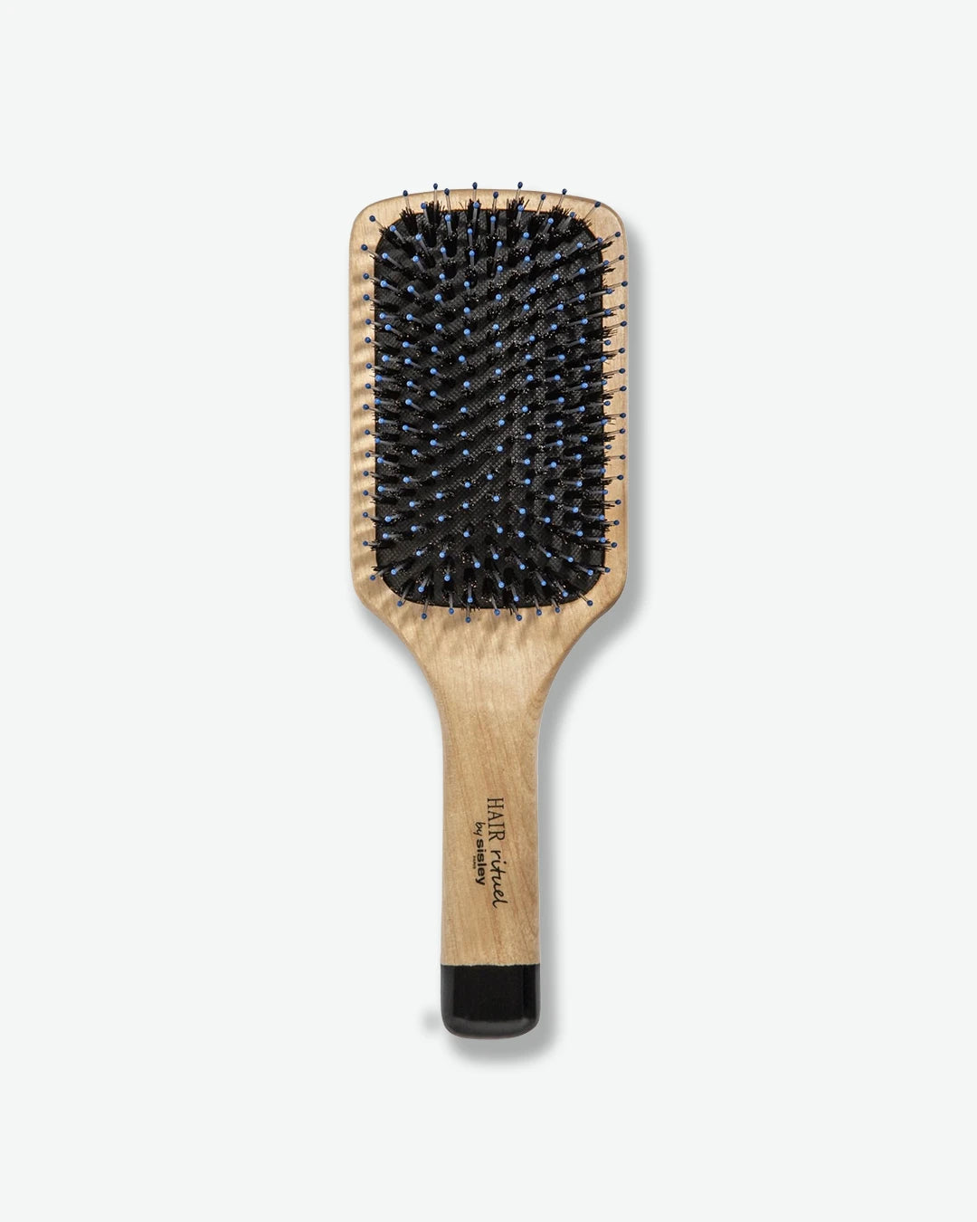 A top-down view of a HAIR Rituel by Sisley paddle hairbrush, featuring a natural light-wood handle and body, a black cushion pad, mixed bristles with bright blue tips, and a glossy black base on the handle.