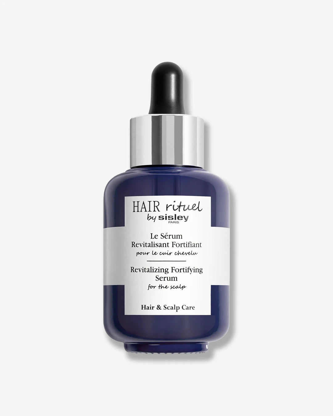 A glossy, deep blue glass dropper bottle of Hair Rituel by Sisley Revitalizing Fortifying Serum for the scalp, featuring a wide white wrap-around label and a polished silver dropper cap.