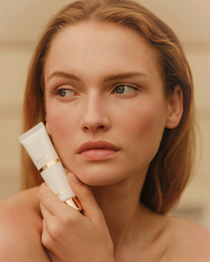 Close-up portrait of a woman with auburn hair resting her chin against a white tube of Sisley Phyto-Teint Primer.