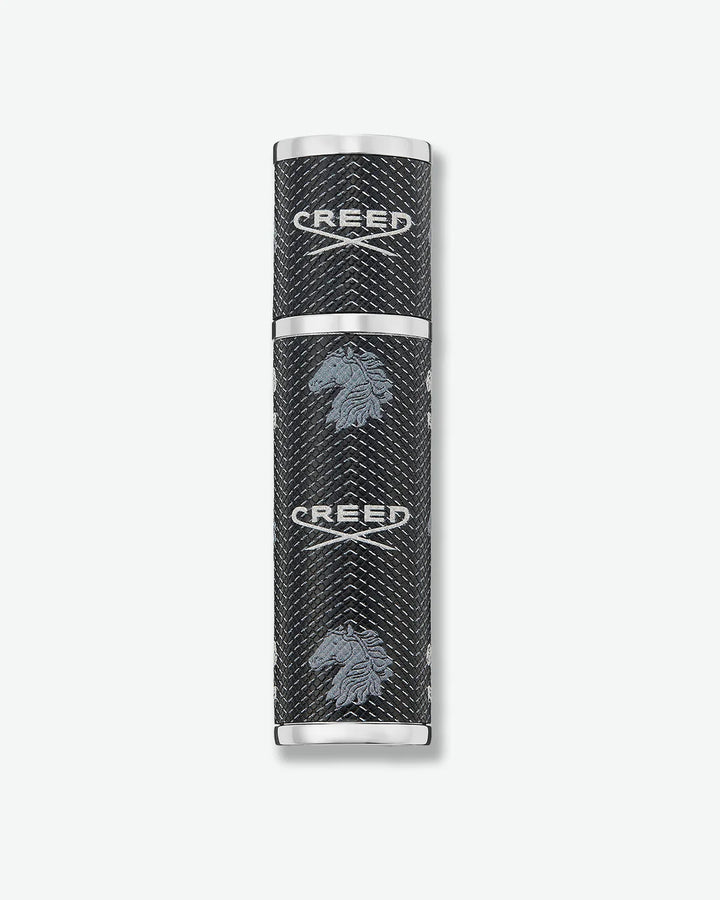 Black cylindrical Creed fragrance travel atomizer with textured leather-like finish, silver-tone metal accents, and repeating Creed logo and horse emblem on a light gray background.