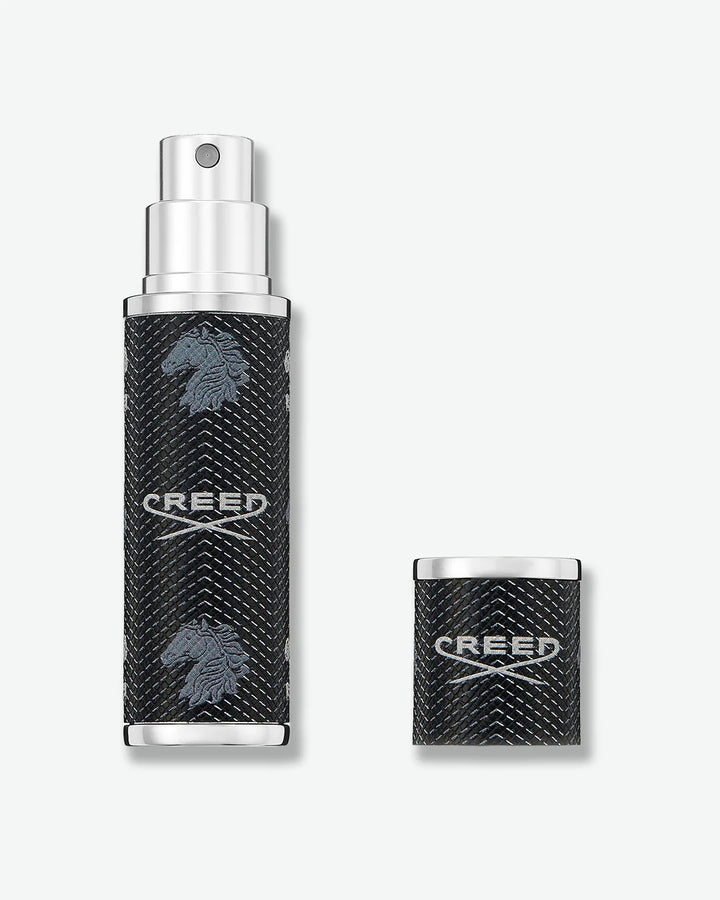 Black cylindrical Creed fragrance travel atomizer with textured leather-like finish, silver-tone metal accents, repeating Creed logo and horse emblem, with cap off, and a silver pump spray nozzle on a light gray background.