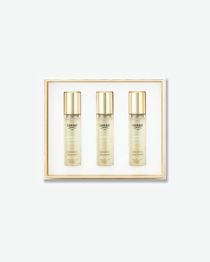 A Creed Aventus trilogy fragrance discovery set showcasing three travel spray vials with gold caps in a white box with a gold inner border, featuring Aventus Cologne, Aventus, and Absolu Aventus, isolated on a light grey background.