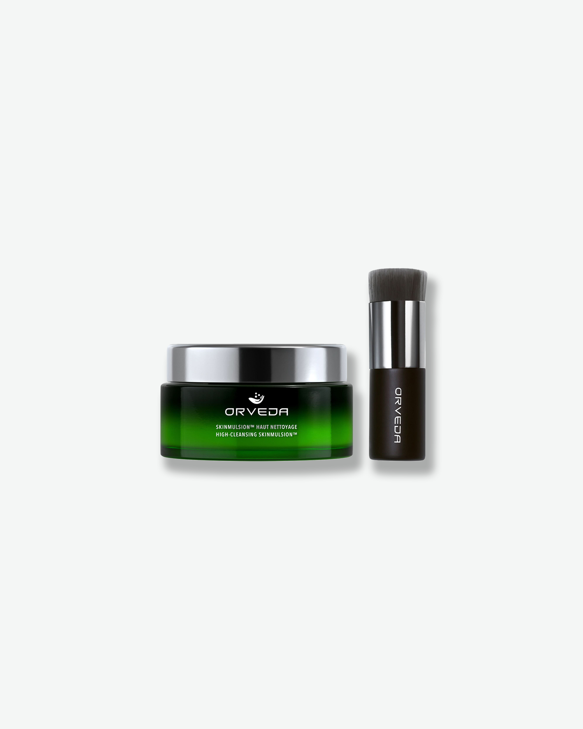 Product shot featuring an Orveda "Skinmulsion™ / High-Cleansing Skinmulsion™" jar standing next to a skincare brush. The low, wide green gradient glass jar with a polished silver cap sits beside a short, dense blending brush with a dark handle, silver ferrule, and charcoal-grey bristles.
