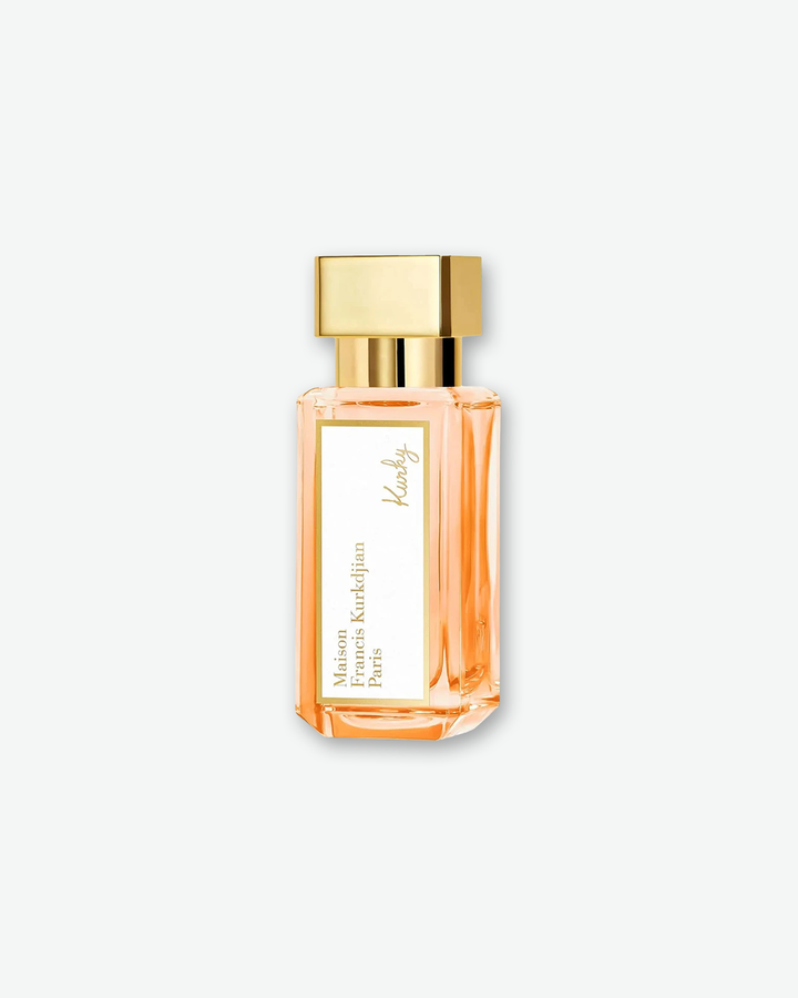 A slightly angled, three-quarter view of a slender, travel-sized Maison Francis Kurkdjian Paris "Kurky" Eau de Parfum bottle. The clear, rectangular glass bottle showcases the peach-tinted liquid inside and is topped with a square gold metallic cap.