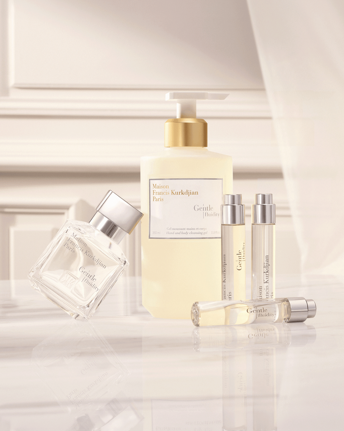 Maison Francis Kurkdjian Gentle Fluidity collection arranged on a reflective surface in a softly lit cream interior, featuring fragrance, body oil, hair perfume, cleansing gel, and travel formats.
