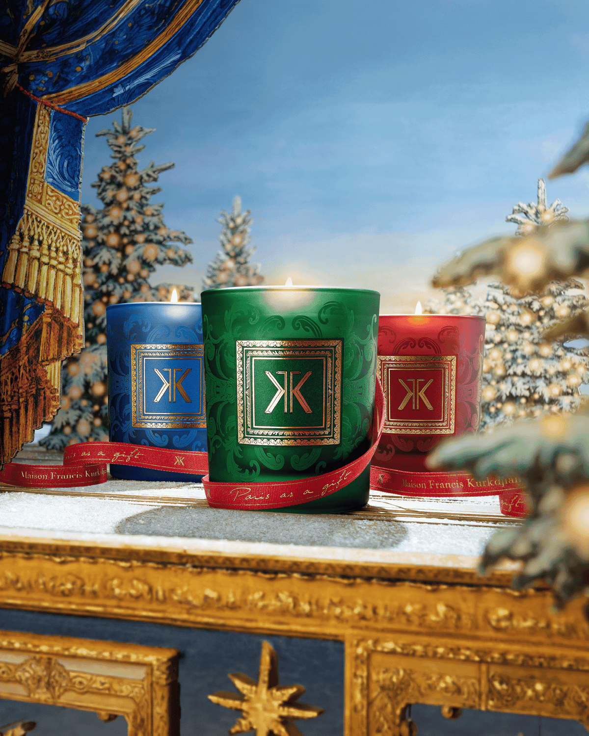 Maison Francis Kurkdjian holiday candles in blue, green, and red displayed in an ornate festive setting with snow-covered Christmas trees, gold detailing, and red ribbon.
