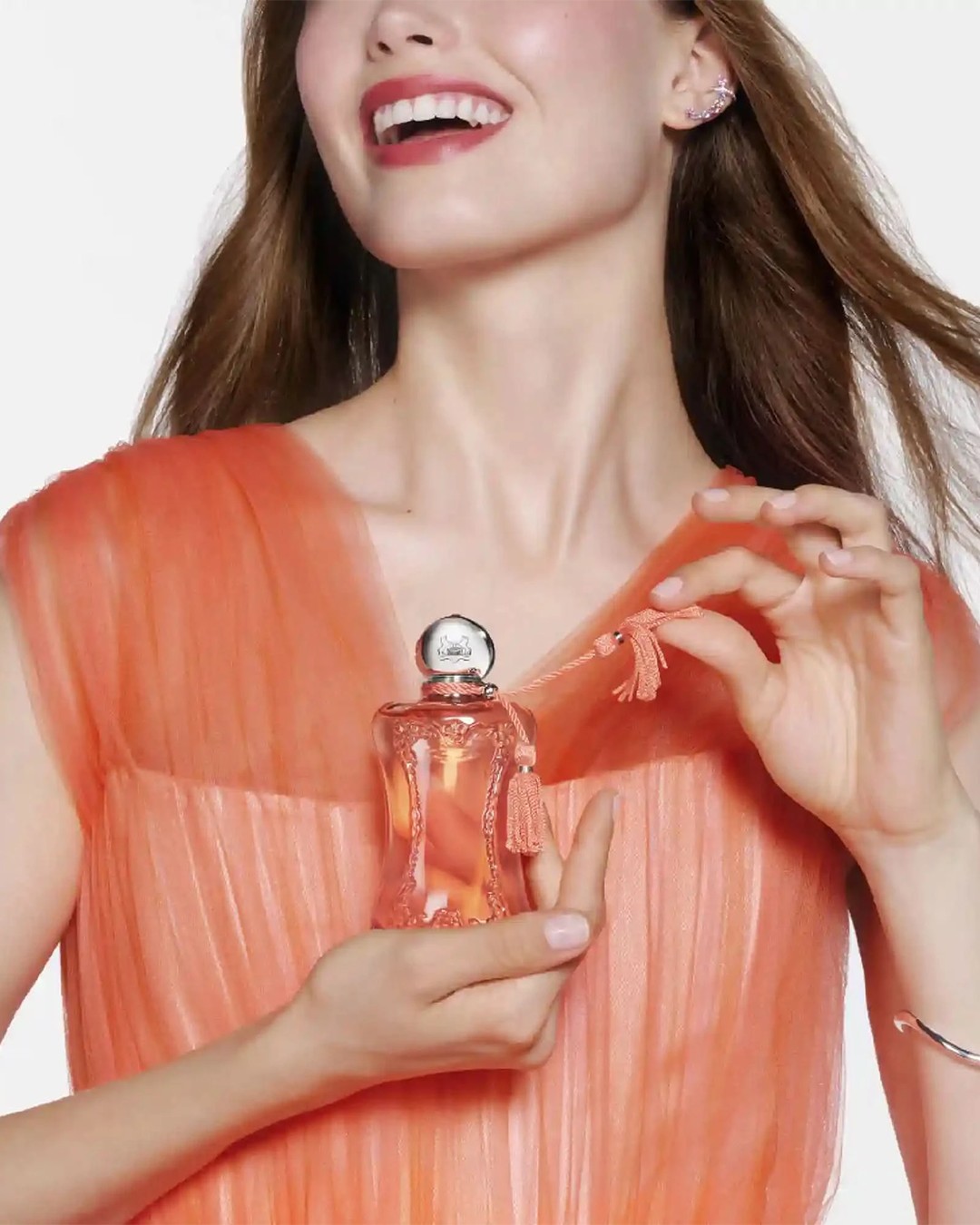 A lifestyle promotional image featuring a woman smiling broadly while holding the peach-colored perfume bottle. She wears an airy, pleated peach and pink tulle dress that matches the tone of the fragrance. Her hands gently cradle the bottle near her chest, with one hand playfully pulling on the twisted tassel accent attached to the silver cap.