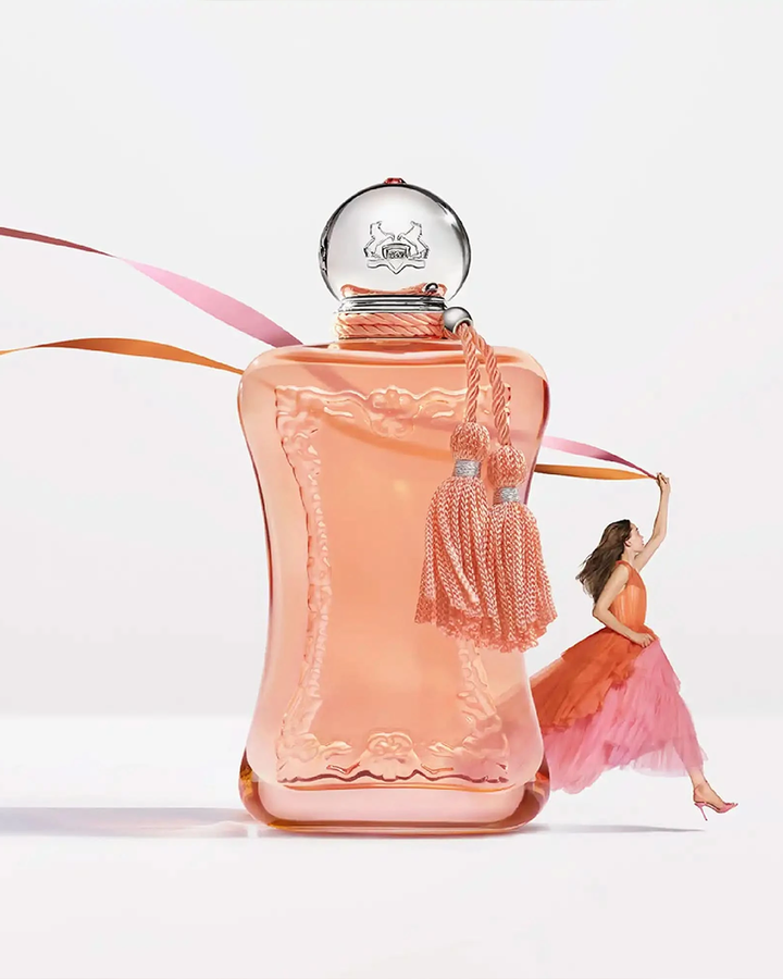 A whimsical, high-fashion campaign graphic showcasing a giant version of the peach fragrance bottle standing on a white surface. In the background, a miniature woman in a flowing peach and pink gown appears to leap through the air, holding a thin pink and orange ribbon that floats gracefully behind the oversized perfume bottle.