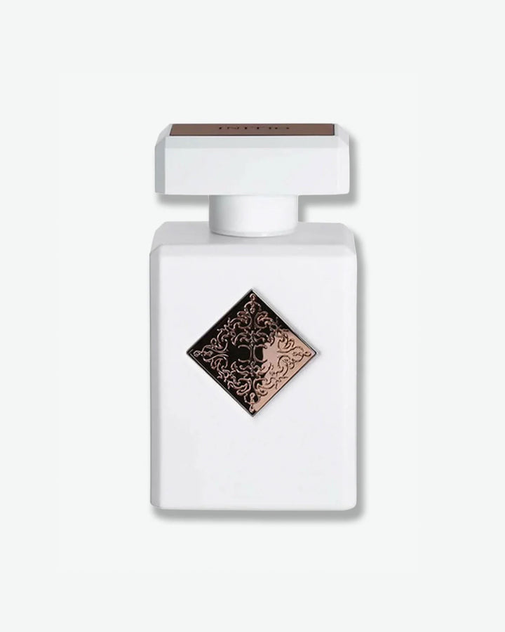 Initio Parfums Privés fragrance in a clean matte white square bottle, featuring a centered rose gold or bronze diamond badge with an intricate engraved filigree emblem, isolated on a light grey background.
