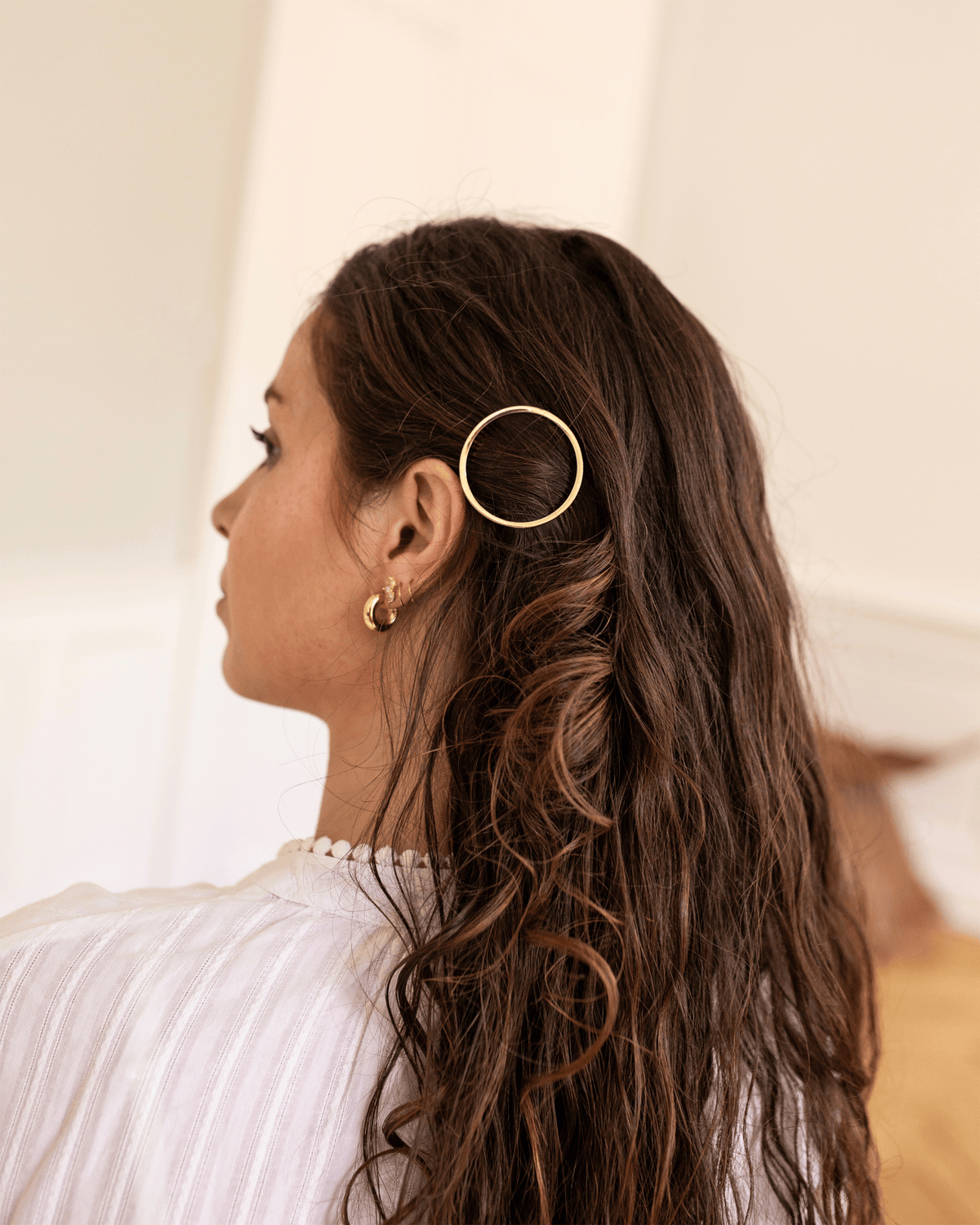 Side view of a model with long, wavy brown hair wearing a circular gold-tone Bachca hair pin above her ear, paired with a white blouse and gold hoop earrings.