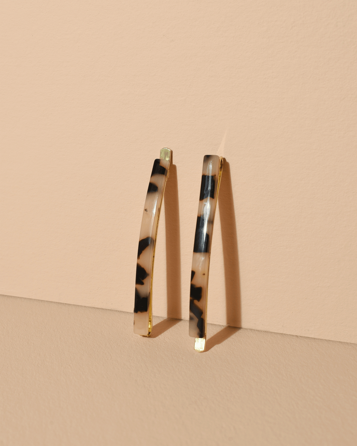 Two slim Bachca Paris tortoiseshell hair clips with gold-tone metal edges, standing upright against a warm beige background.