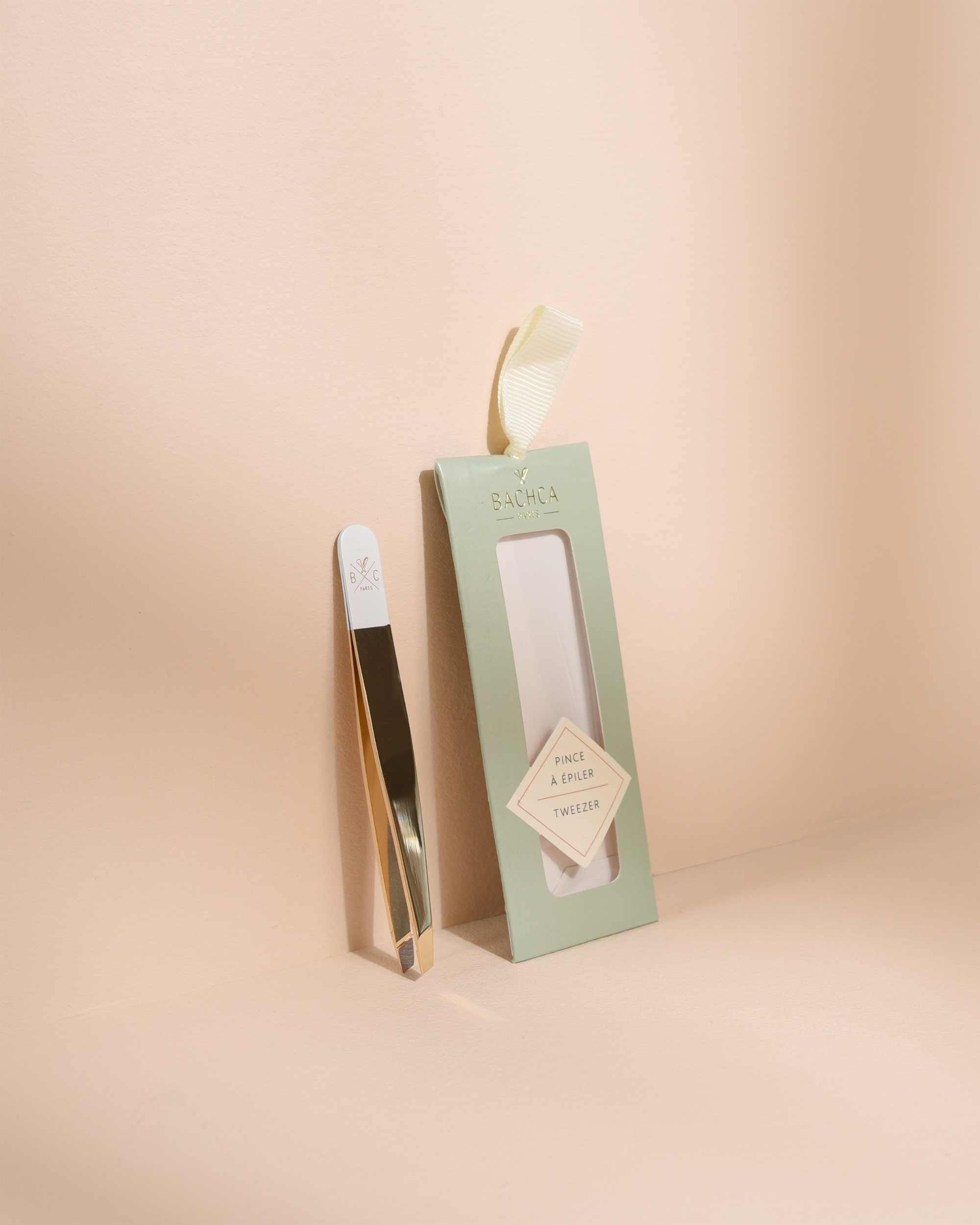 Luxury gold and white slanted tweezers standing upright next to their empty sage green packaging box on a warm beige background.