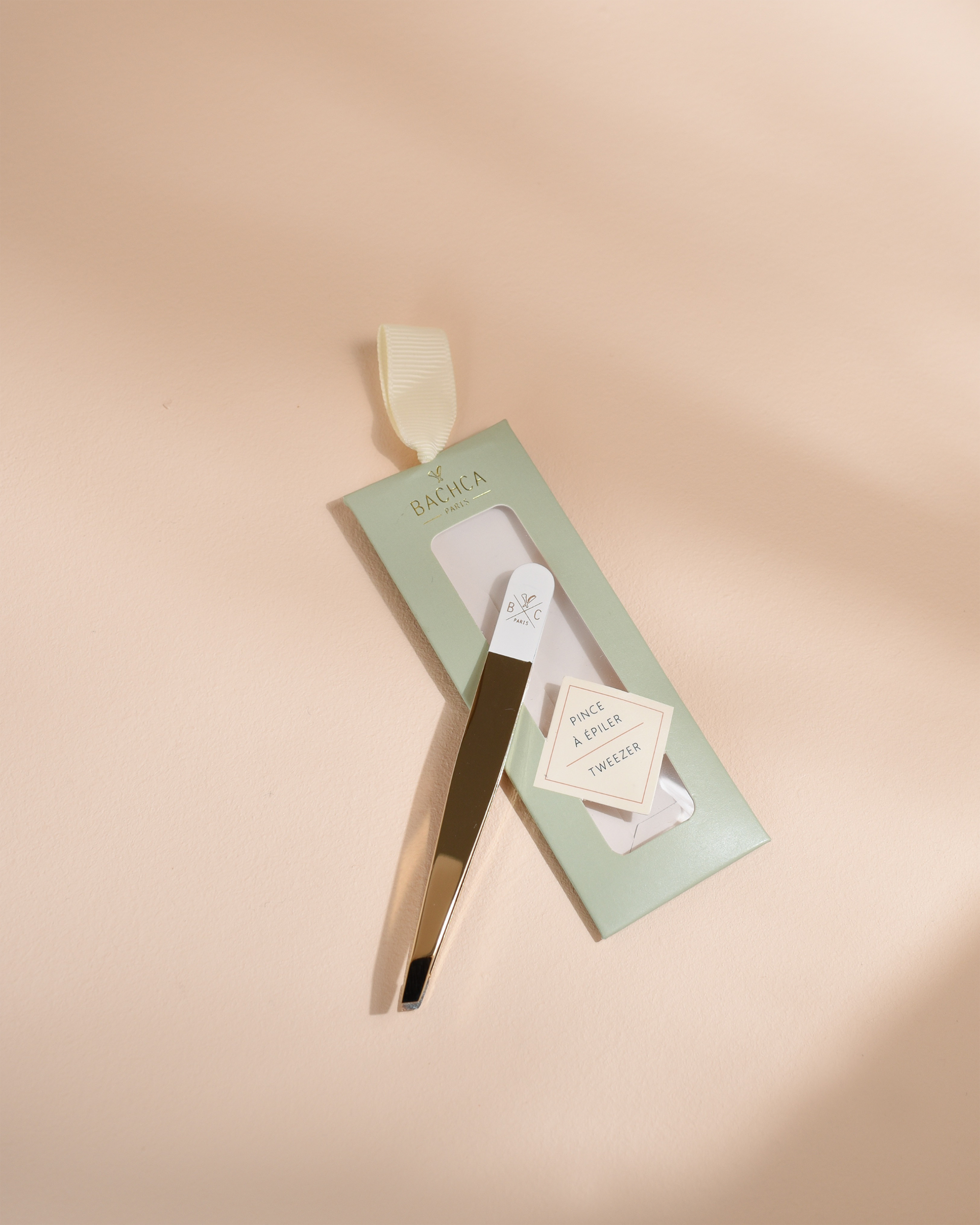 Gold and white slanted tweezers leaning against their empty sage green product box on a beige surface