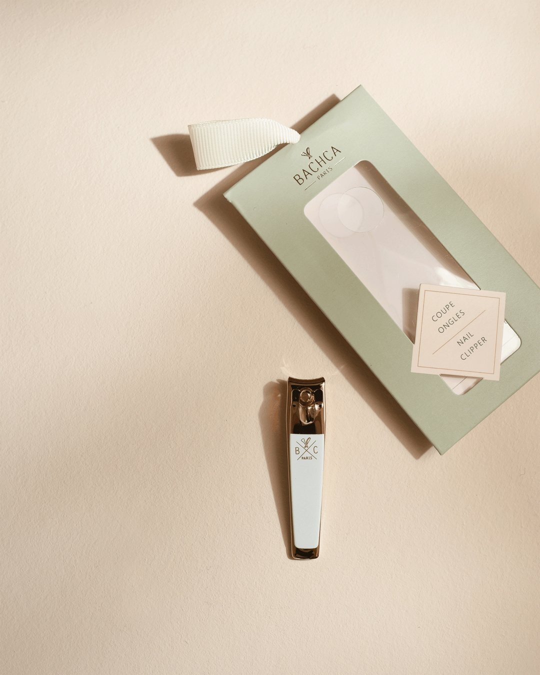 Gold and white nail clipper displayed next to its empty sage green box on a neutral, warm beige surface with elegant shadows.